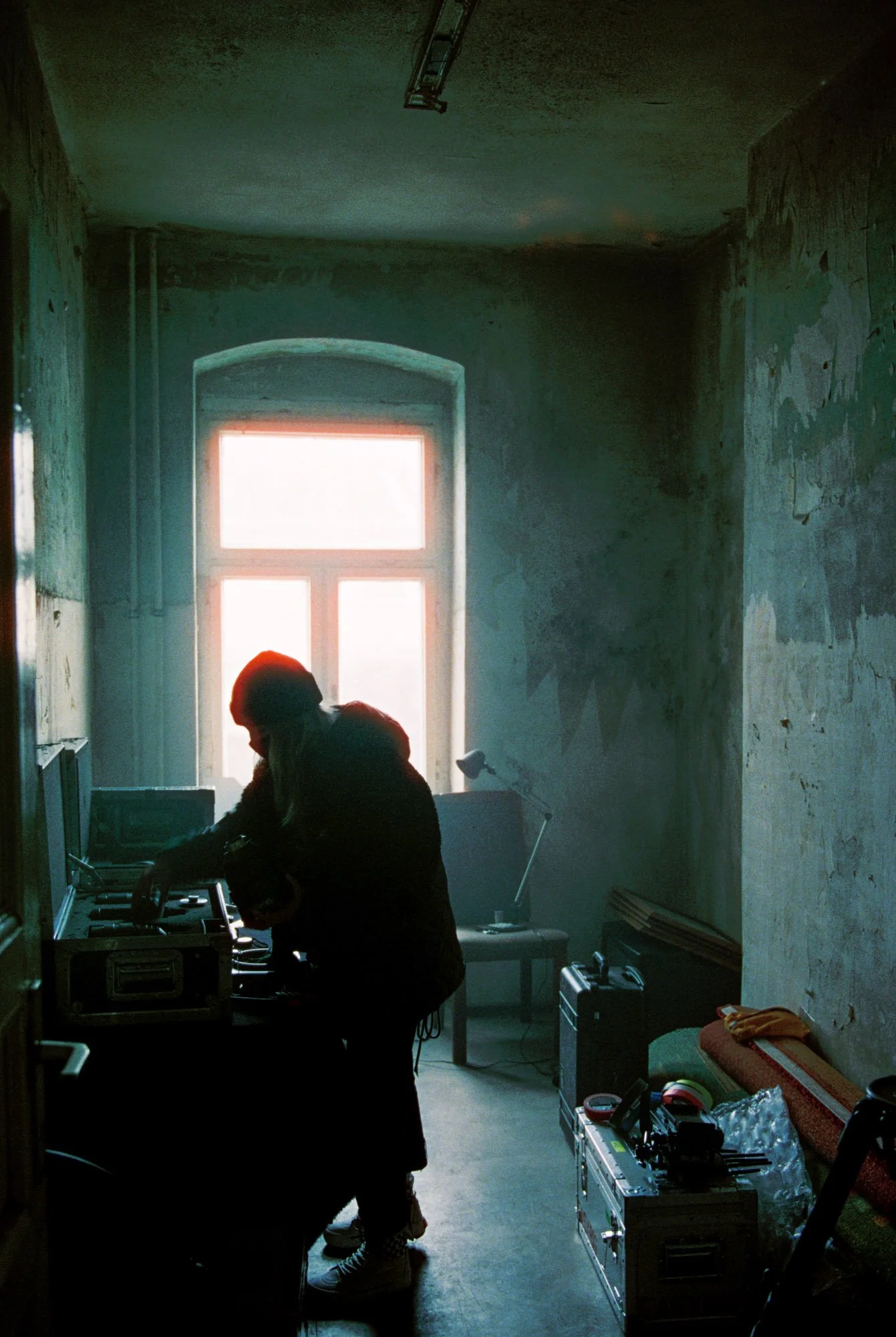 A person is working with DJ equipment in a dimly lit room with a large window and gritty, textured walls.
