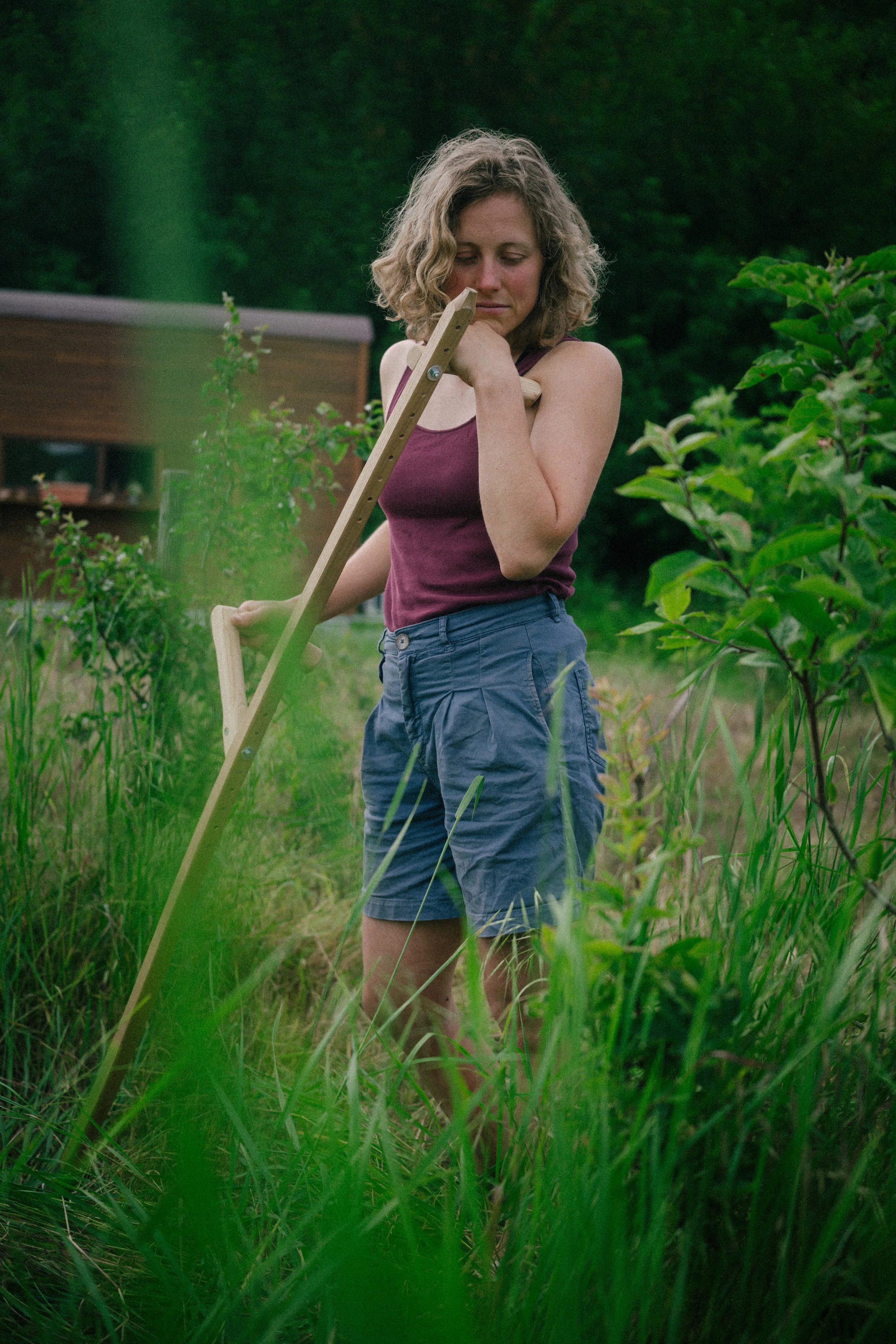 A young woman with curly blonde hair, wearing a purple sleeveless top and blue shorts, standing in a lush garden or field surrounded by green plants. She is holding a wooden gardening tool, possibly a rake, over her shoulder and looking down thoughtf