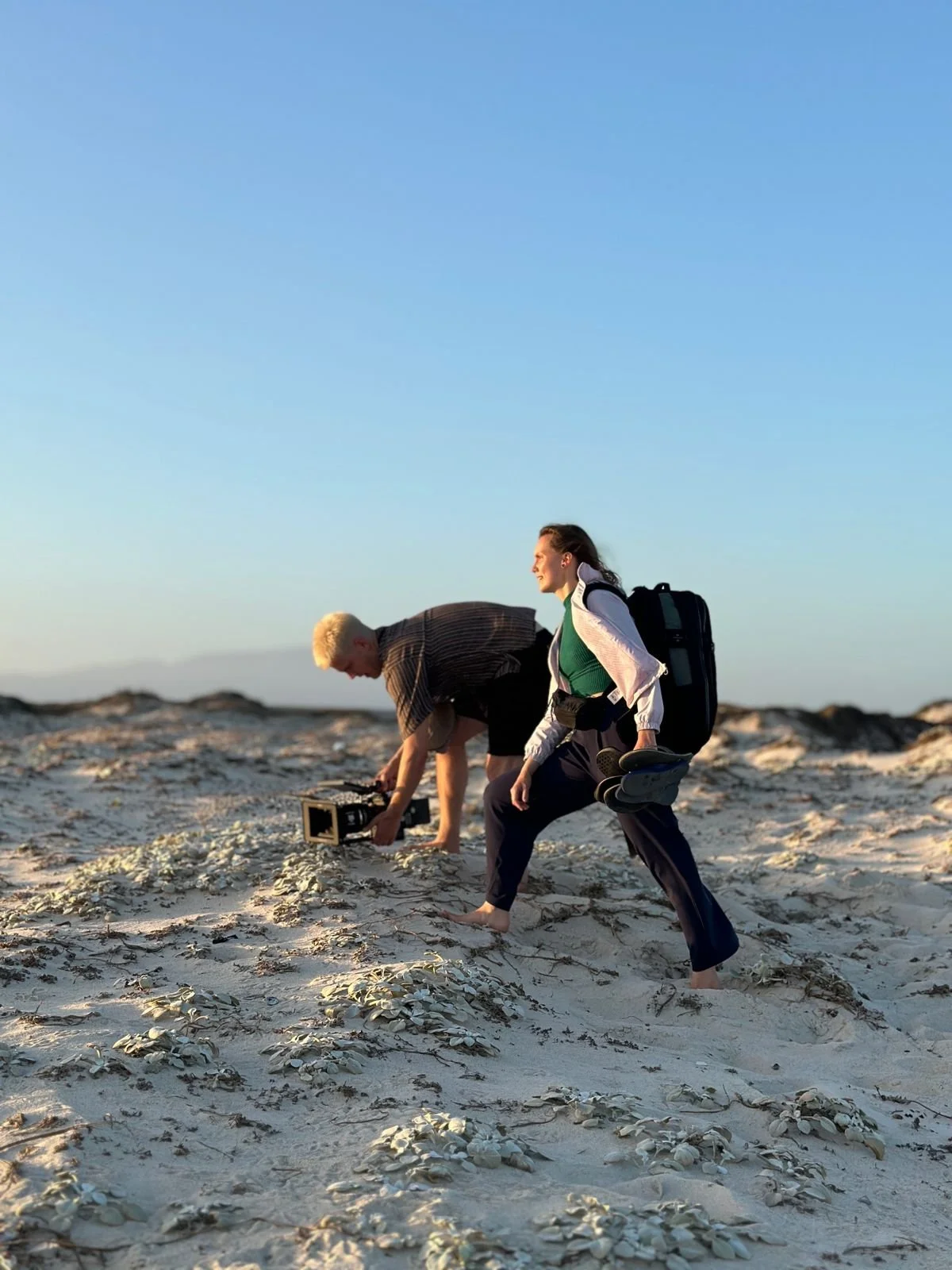Two people, a man and a woman, walking barefoot on a sandy beach with shells, under a clear blue sky. The woman carries a backpack and holds shoes, while the man is kneeling and using a camera.