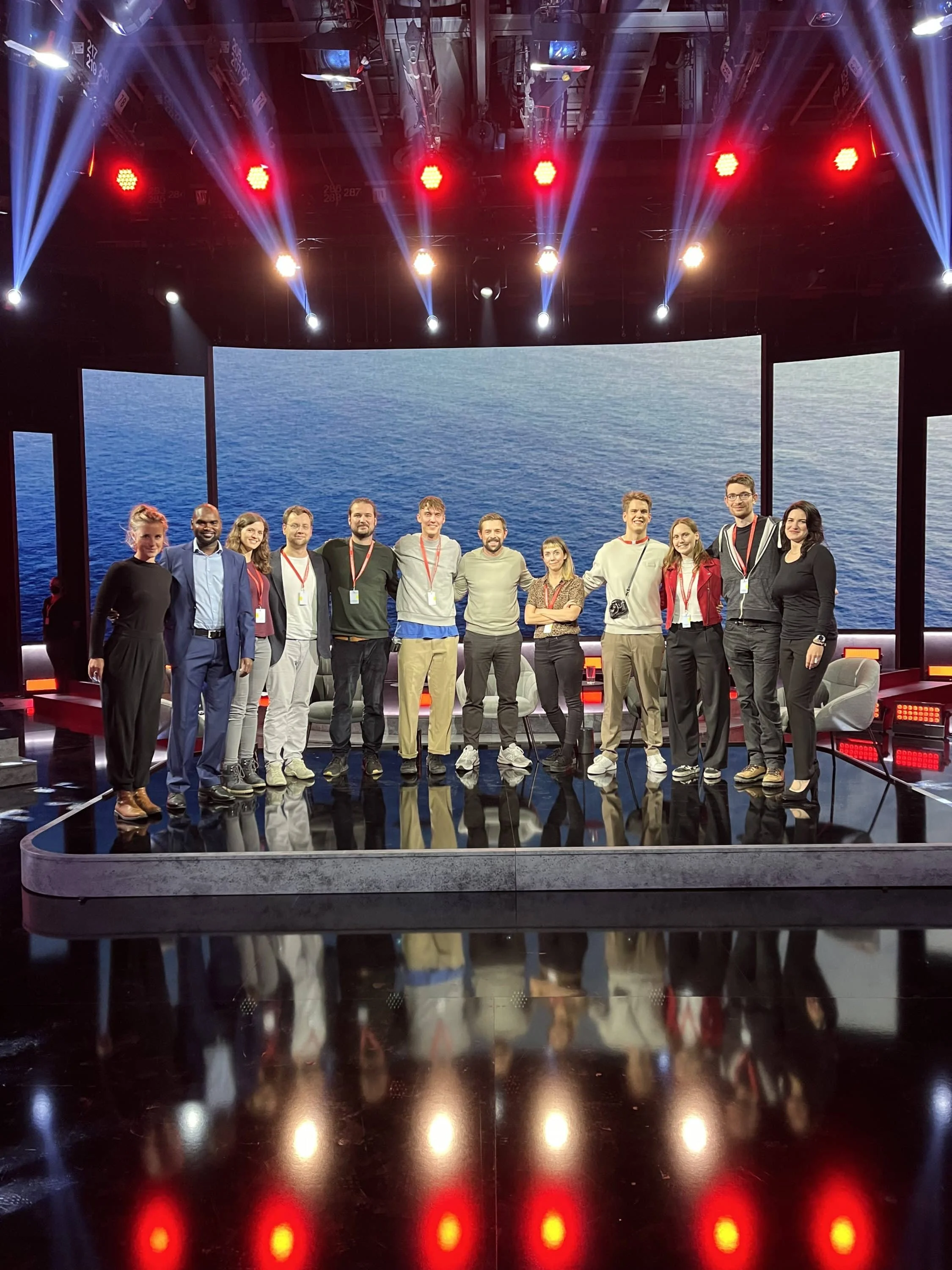Group of 12 people standing on a stage with a large digital screen behind them displaying an ocean scene.
