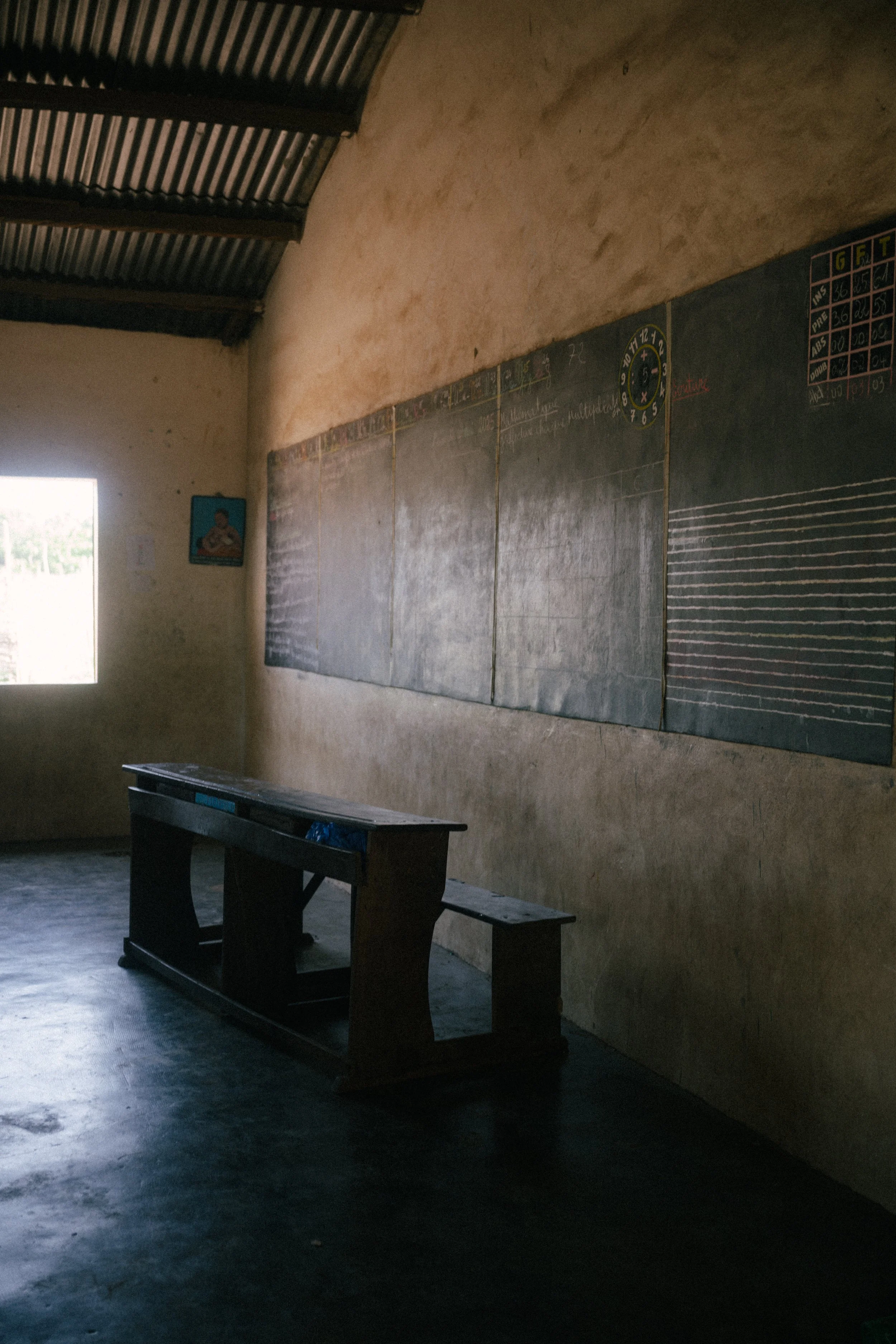 An empty classroom with a large blackboard on the wall, a wooden desk and bench, a small picture on the wall, and a window letting in natural light.
