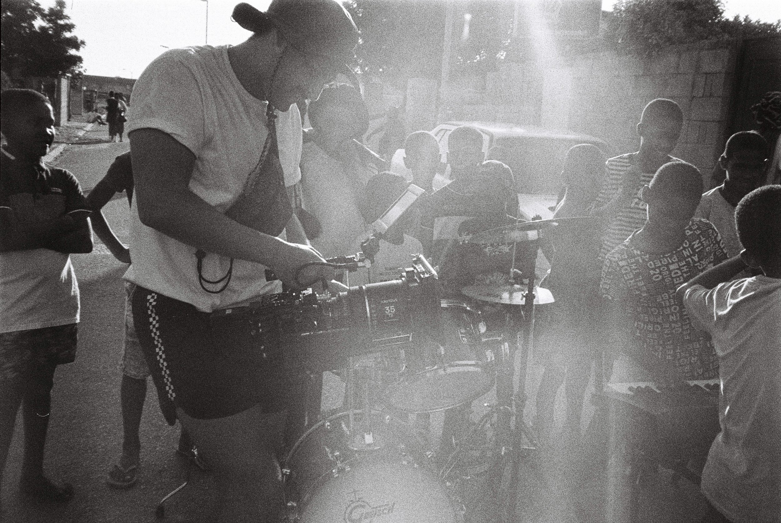 A woman operating a camera surrounded by a group of children and adults outdoors in sunlight.