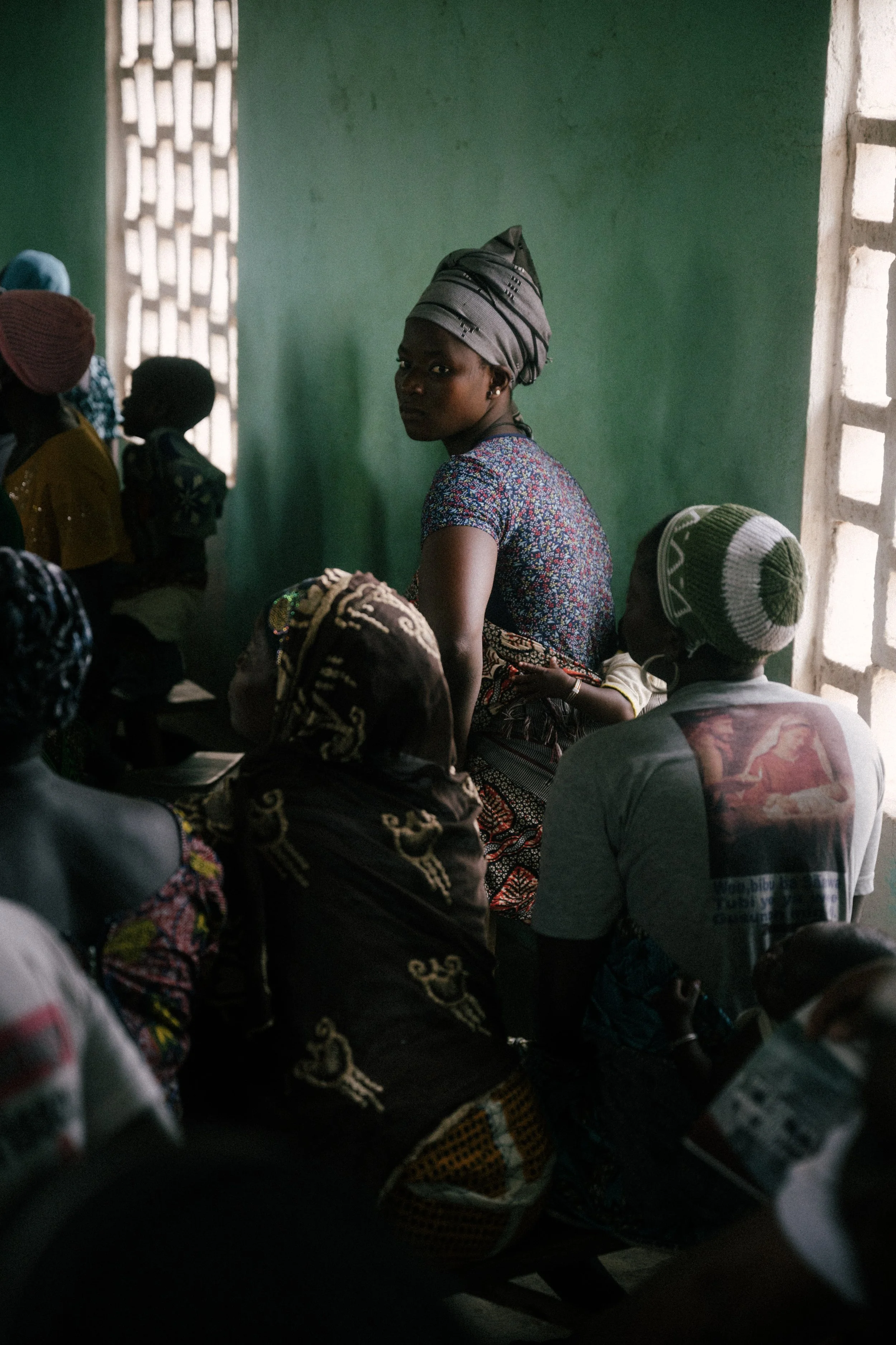 A group of women and girls sitting closely together inside a room with green walls and patterned clothing, some wearing headscarves, with sunlight coming in through a window on the right.