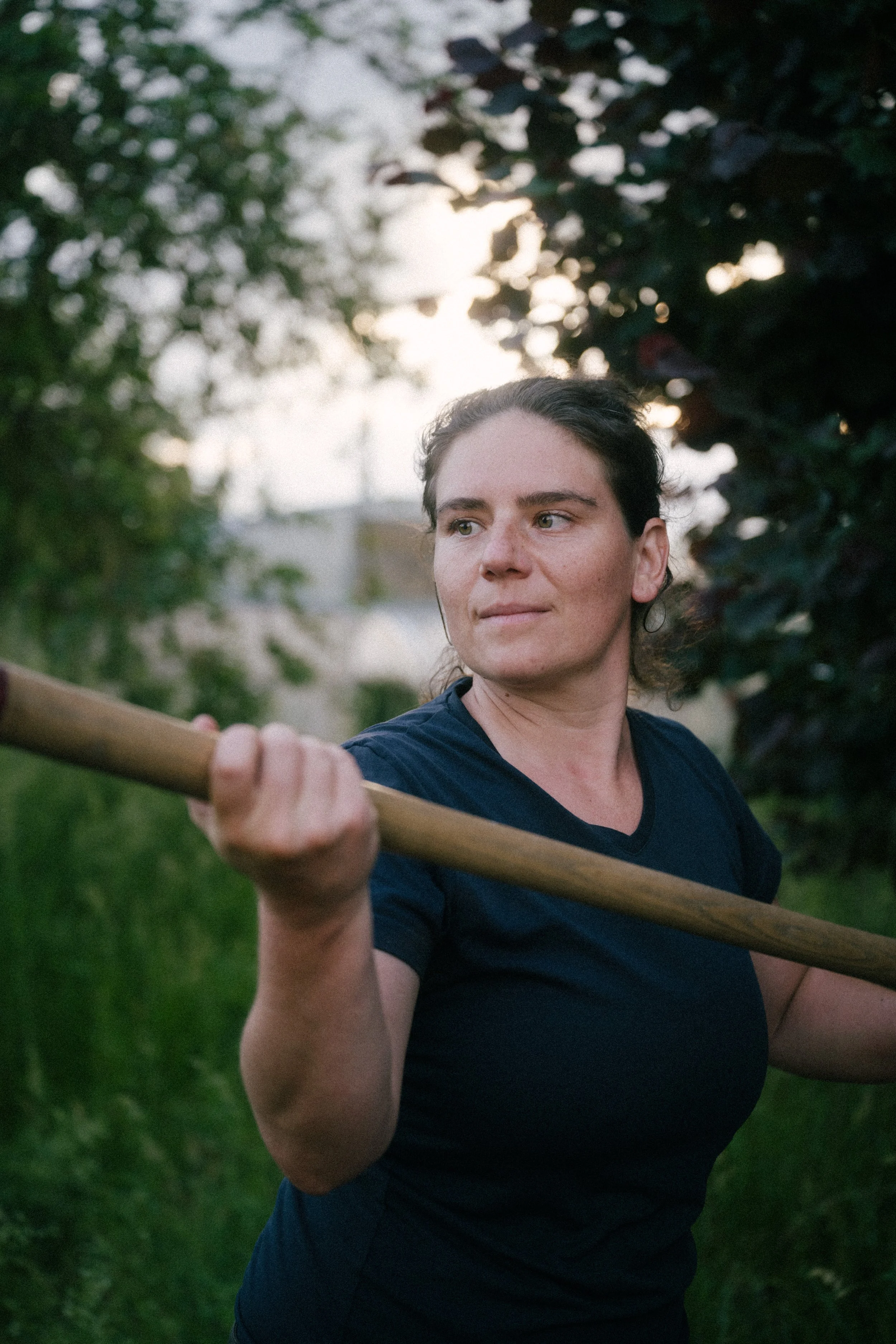 A woman with dark hair pulled back, wearing a navy blue shirt, holds a long wooden stick across her shoulders. She stands outdoors in a green, leafy area with blurred trees and sunlight in the background.