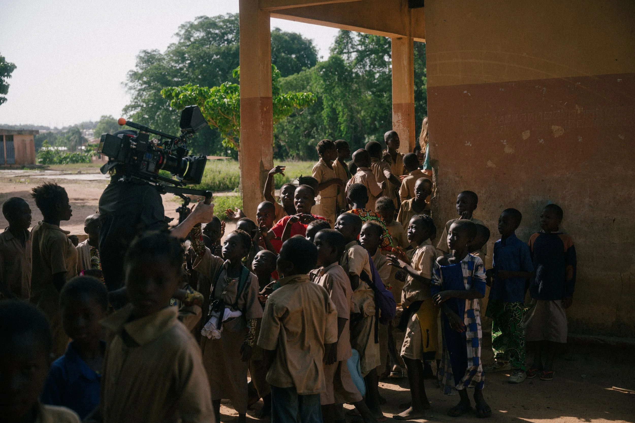 A group of children, some wearing school uniforms, gathered outside a building, with a person filming them with a professional video camera.