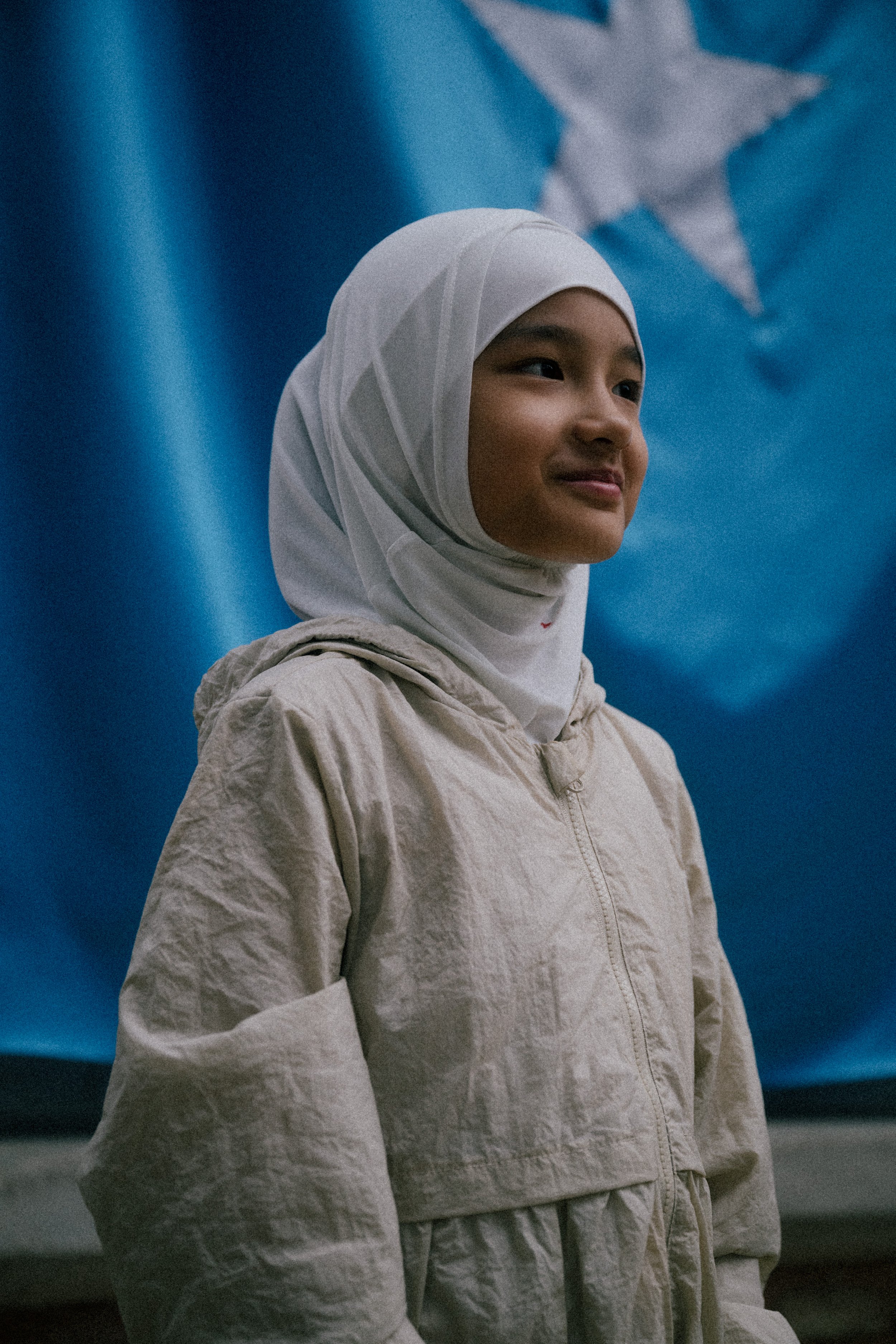 A young girl wearing a white hijab and a beige jacket standing in front of a blue background with a star design.