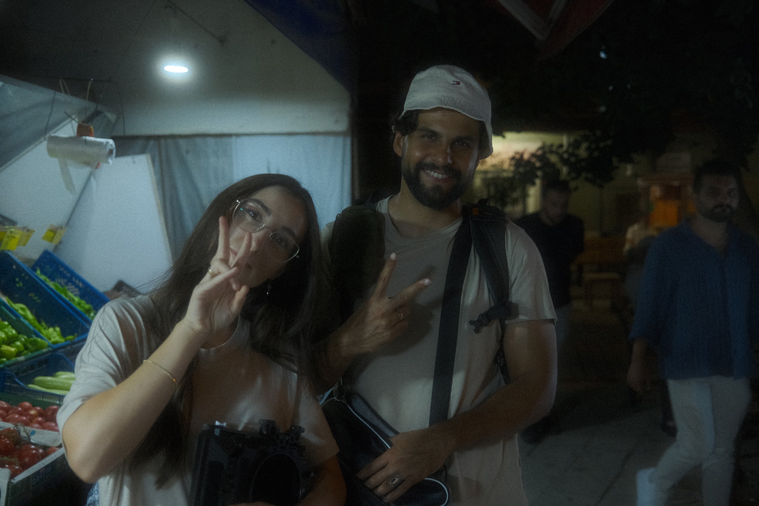 Two people standing together at night, smiling and showing peace signs. The woman on the left is wearing glasses and has long dark hair. The man on the right has facial hair, is wearing a bucket hat, a light-colored T-shirt, and has a camera hanging 