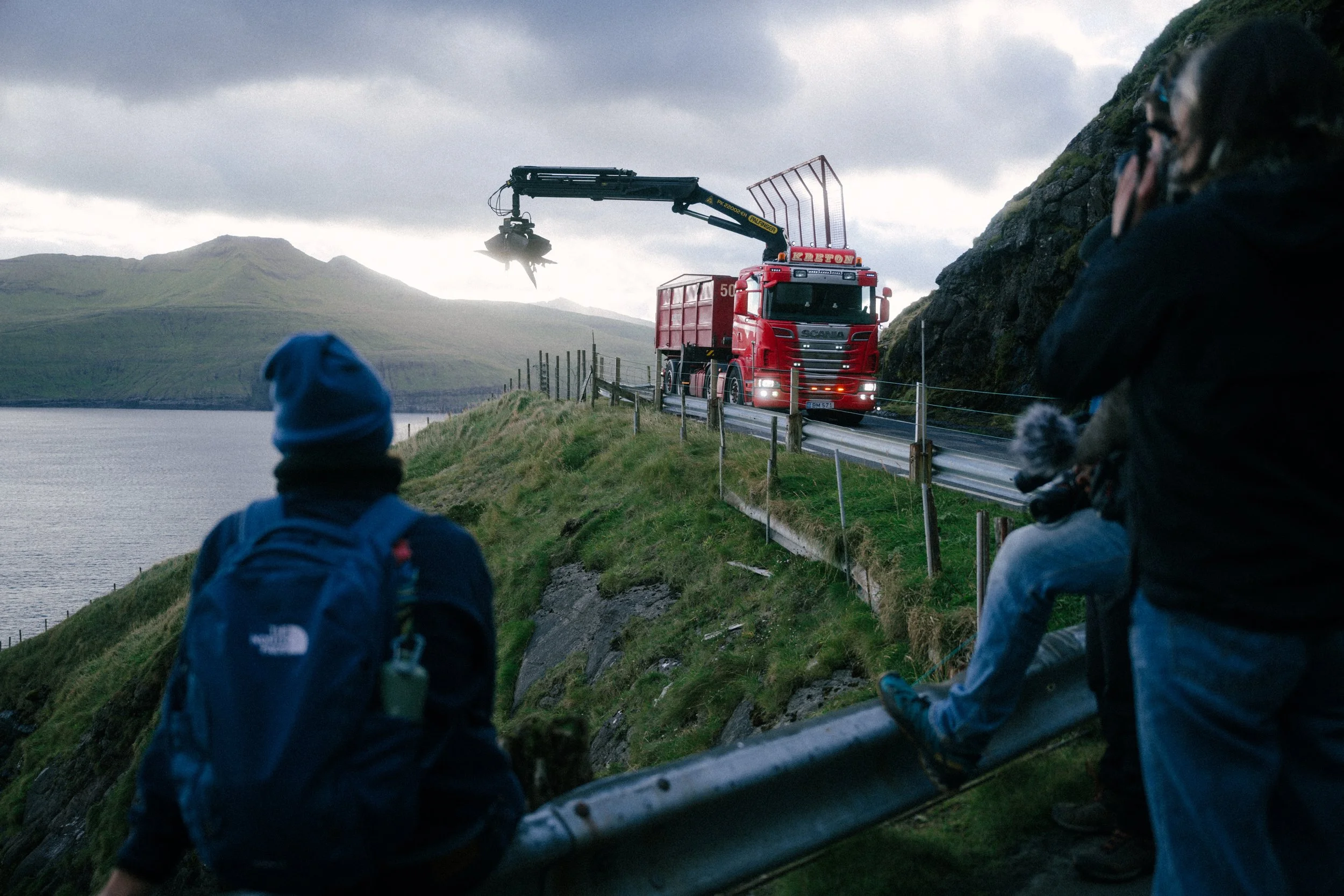 People watching a fire truck with a crane on a mountain road near a body of water, with cloudy skies and green hills in the background.
