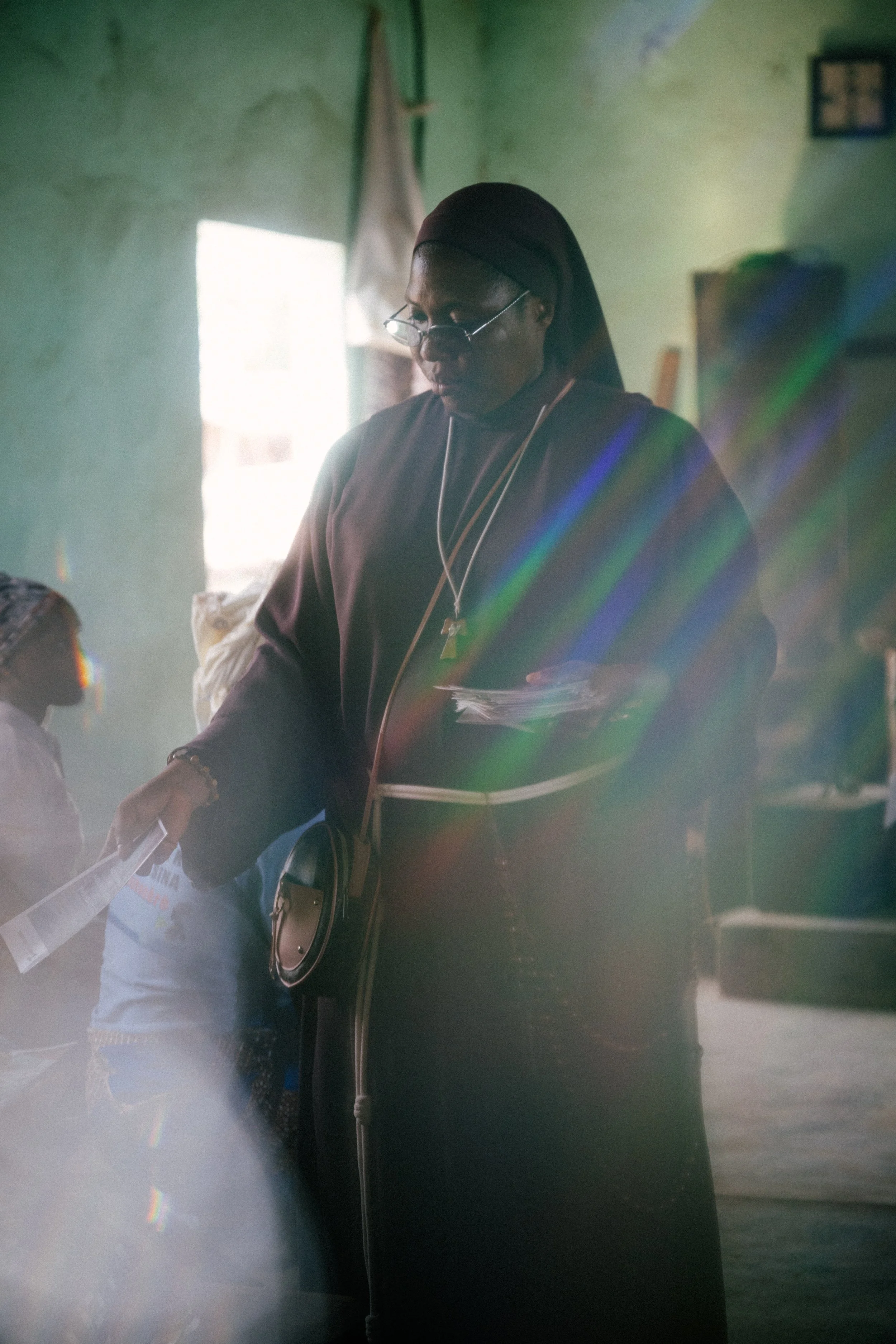 A woman in dark clothing, glasses, and a head covering, standing inside a room with green walls, handing a paper to a person outside the frame.