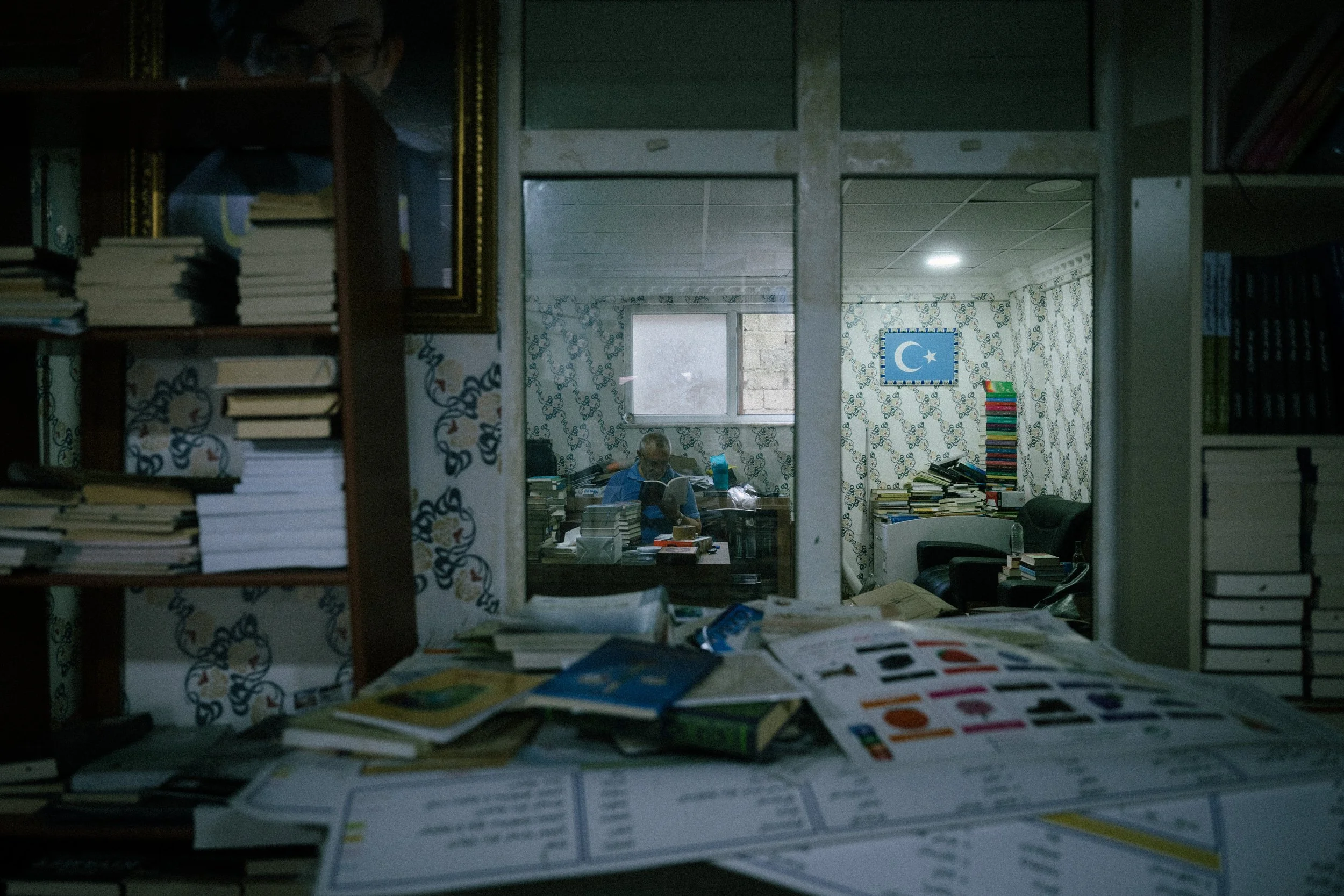 View through a doorway into a room filled with books and papers, with a person sitting at a desk reading.