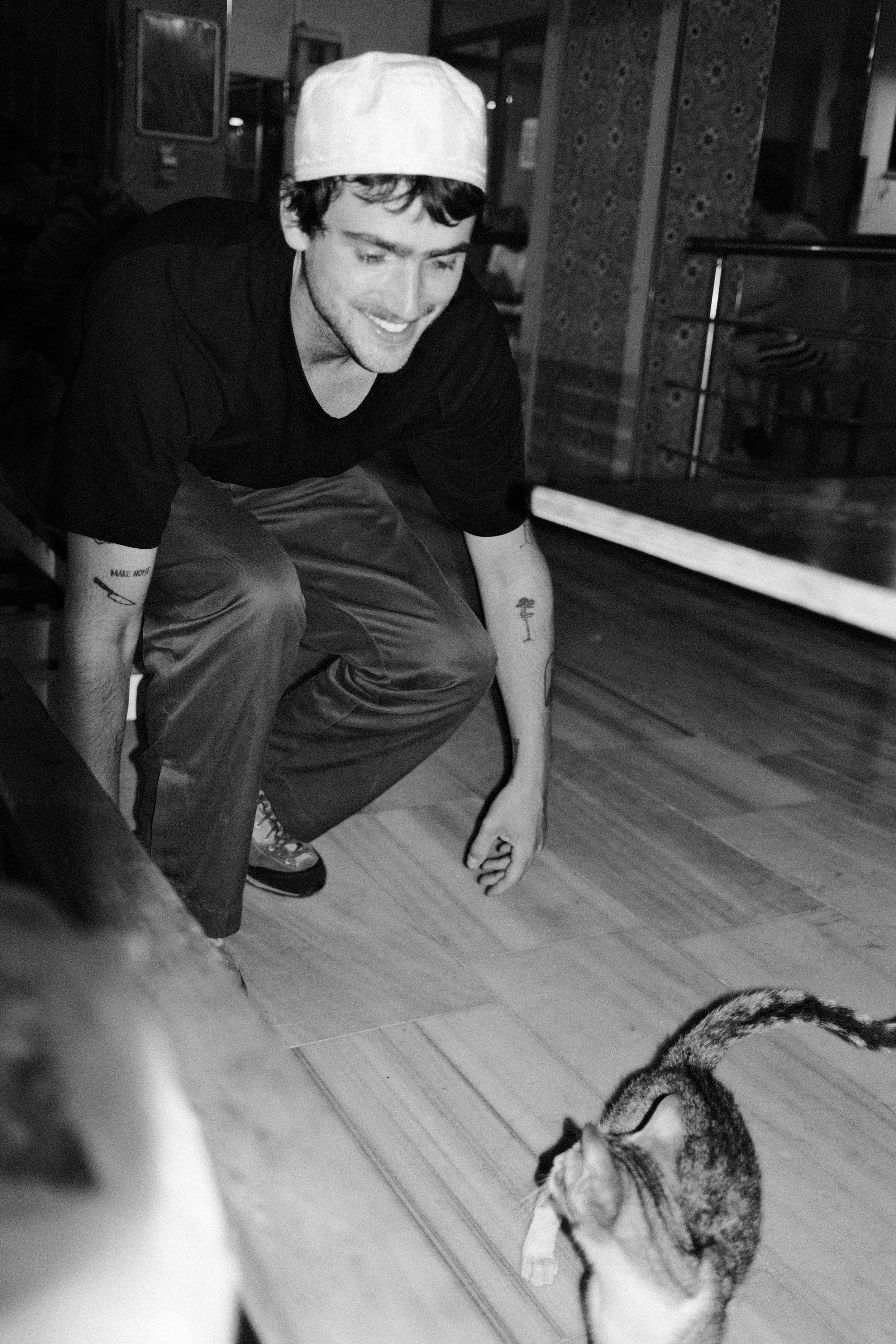 A man wearing a chef's hat crouches down, smiling at a cat sitting on the wooden floor, with another cat partially visible in the foreground.