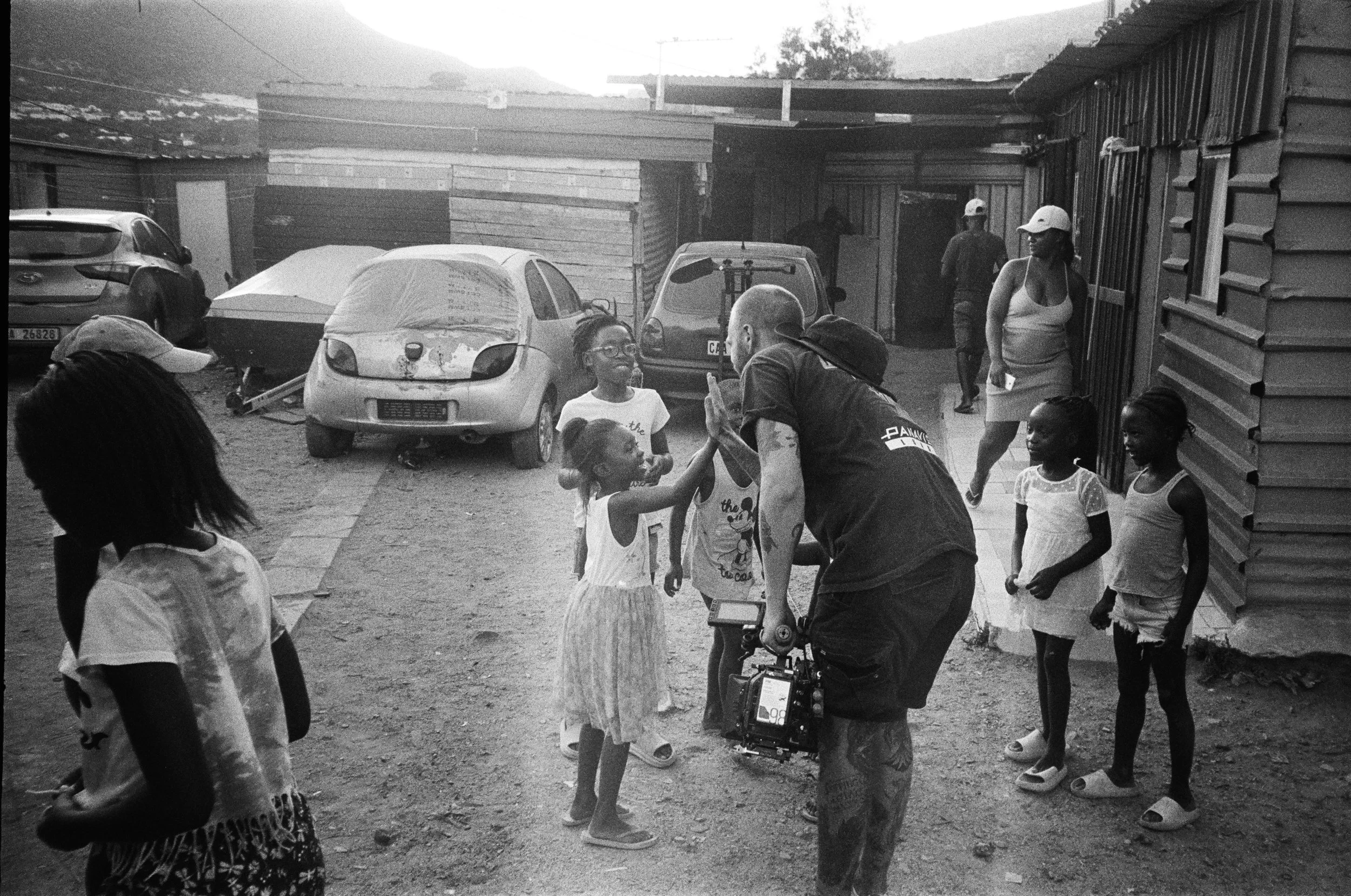 A man takes photos of smiling children interacting outdoors near parked cars and wooden structures in a community setting.