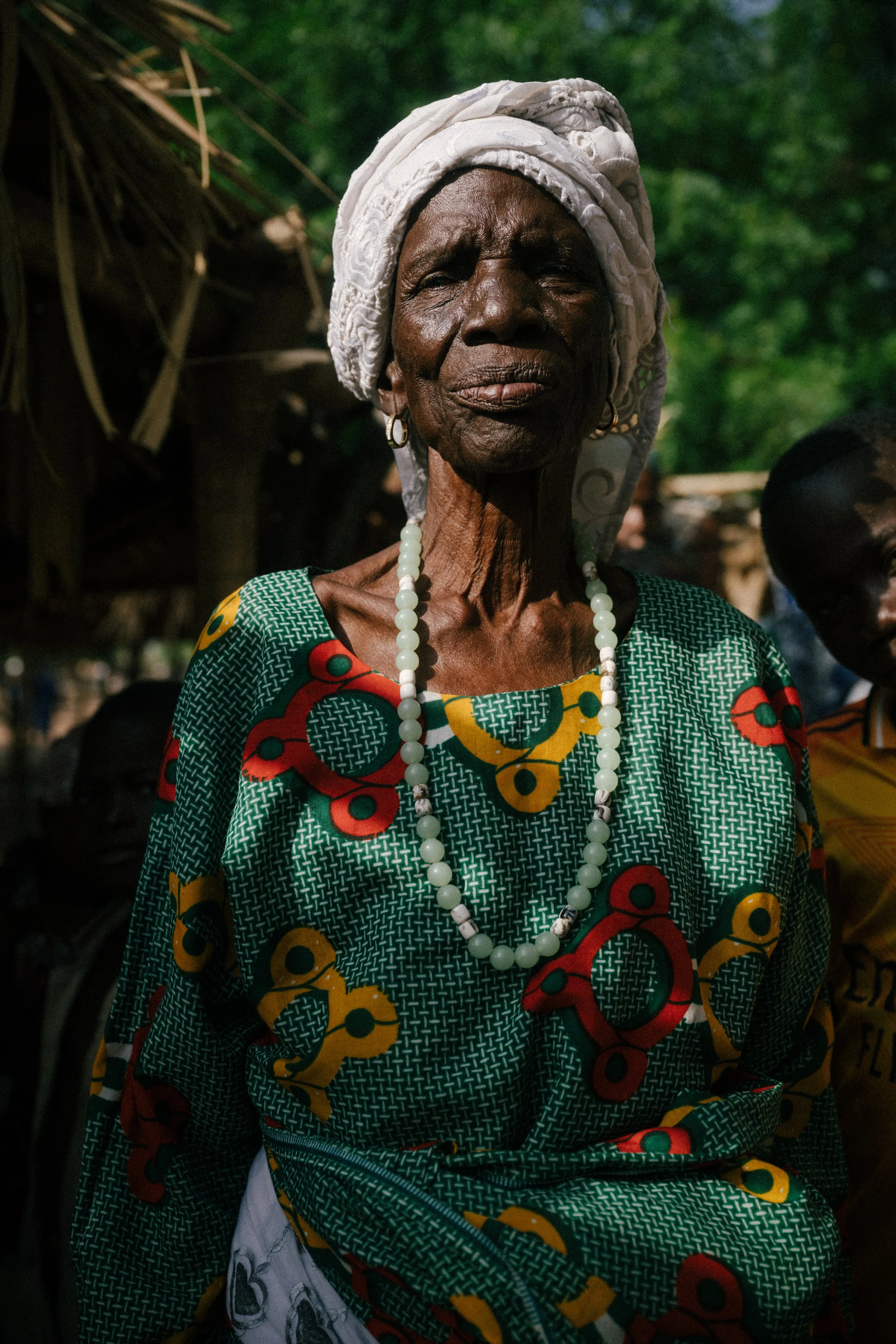 An elderly African woman with dark skin, wearing a white headwrap and earrings, dressed in a colorful traditional green dress with red, yellow, and black patterns, and a white beaded necklace, standing outdoors with green foliage in the background.