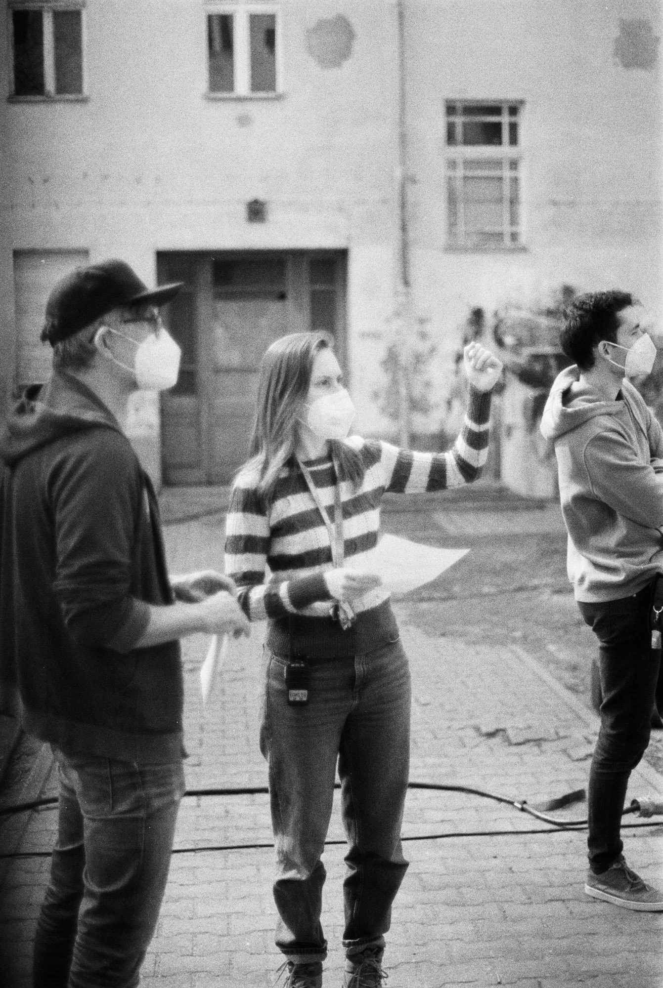 Three people wearing masks standing outdoors during daytime, one woman speaking and gesturing with her fist raised.