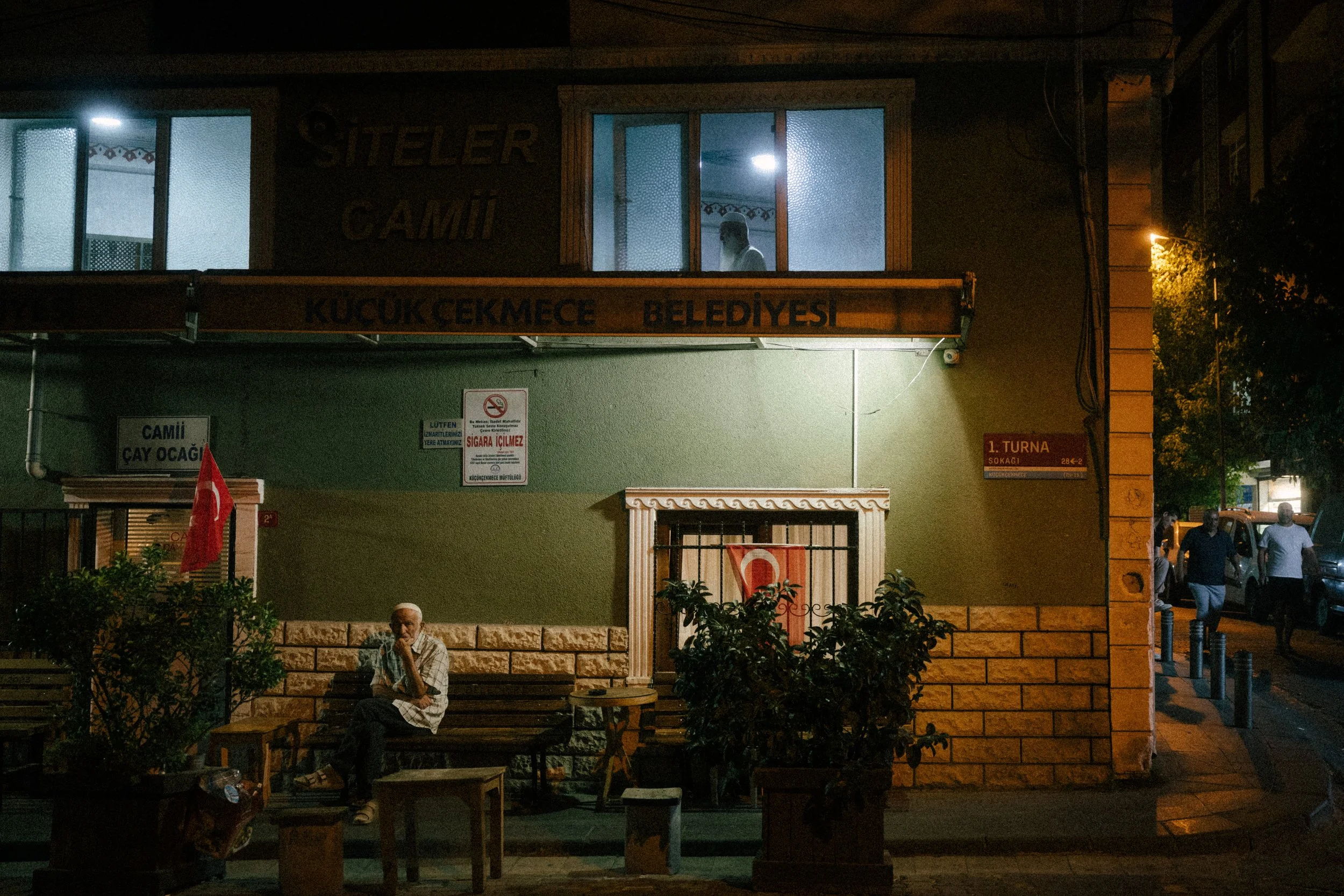 Nighttime scene of a small mosque with a person sitting on a bench in front, Turkish flags, and a street with a few pedestrians.