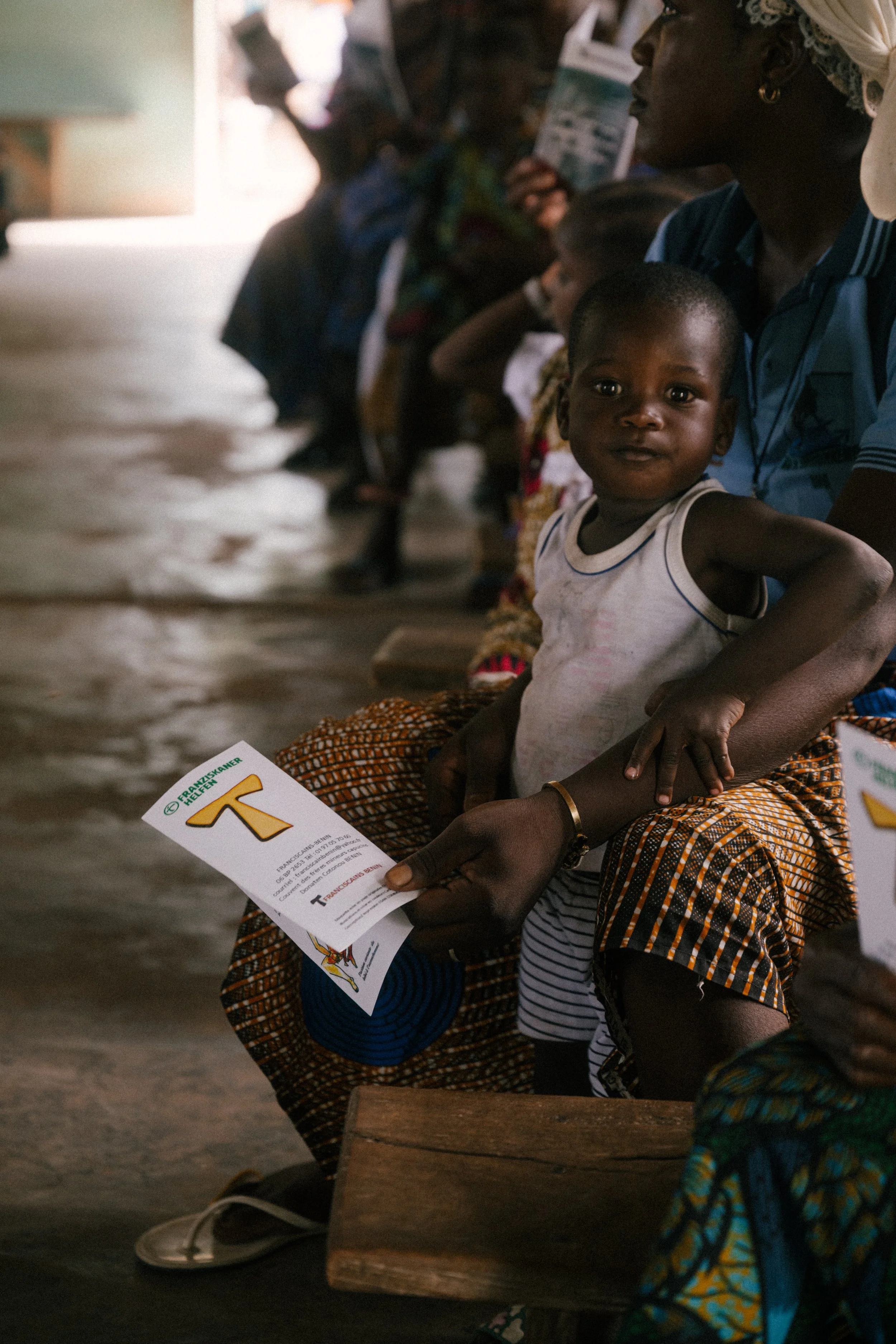 A young girl sitting on a wooden bench in a group, holding a pamphlet, with an adult woman behind her. Other children and adults are visible in the background, waiting or seated.