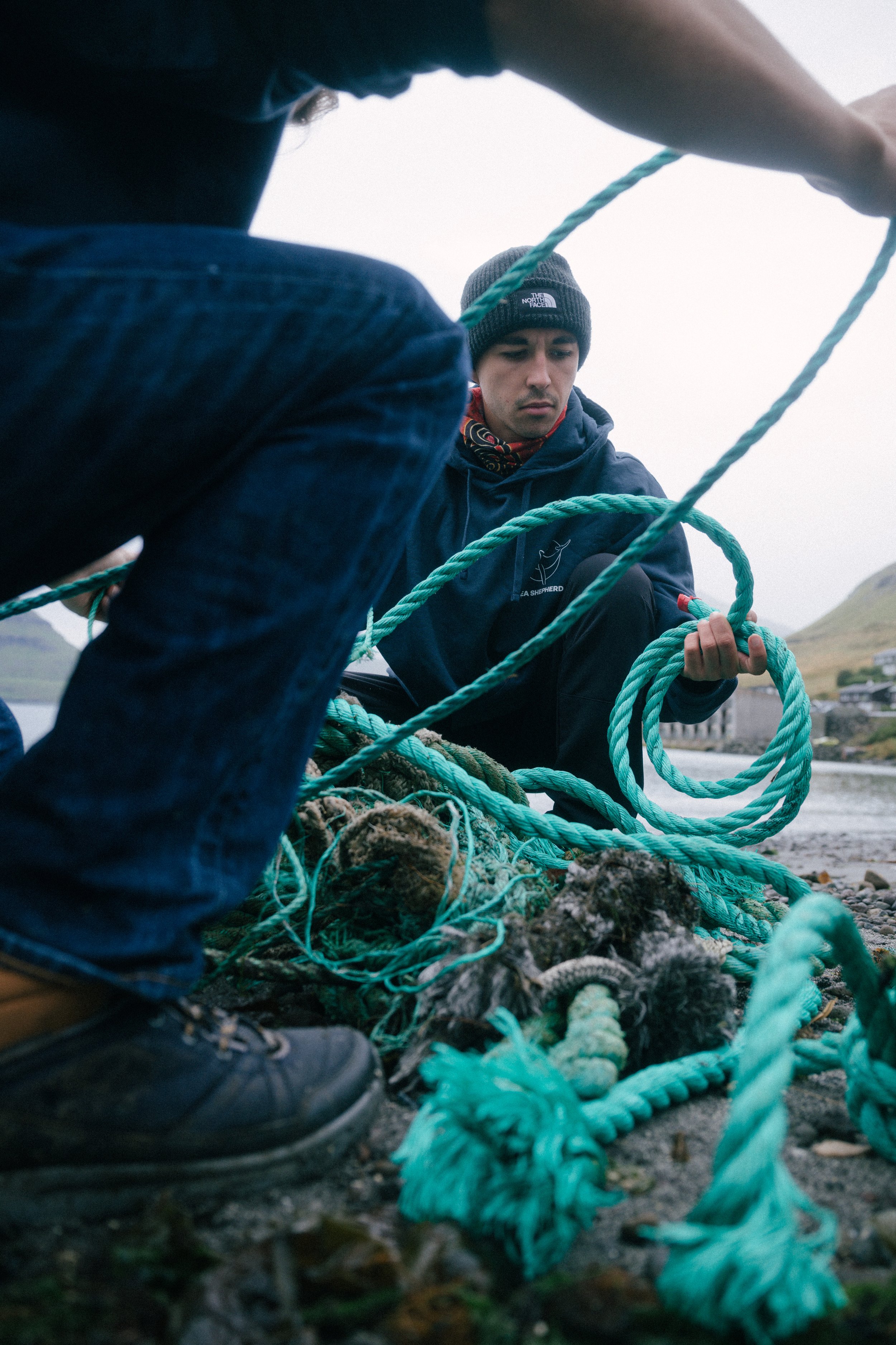 Two people working on tangled ropes on a rocky shoreline with hills and buildings in the background.