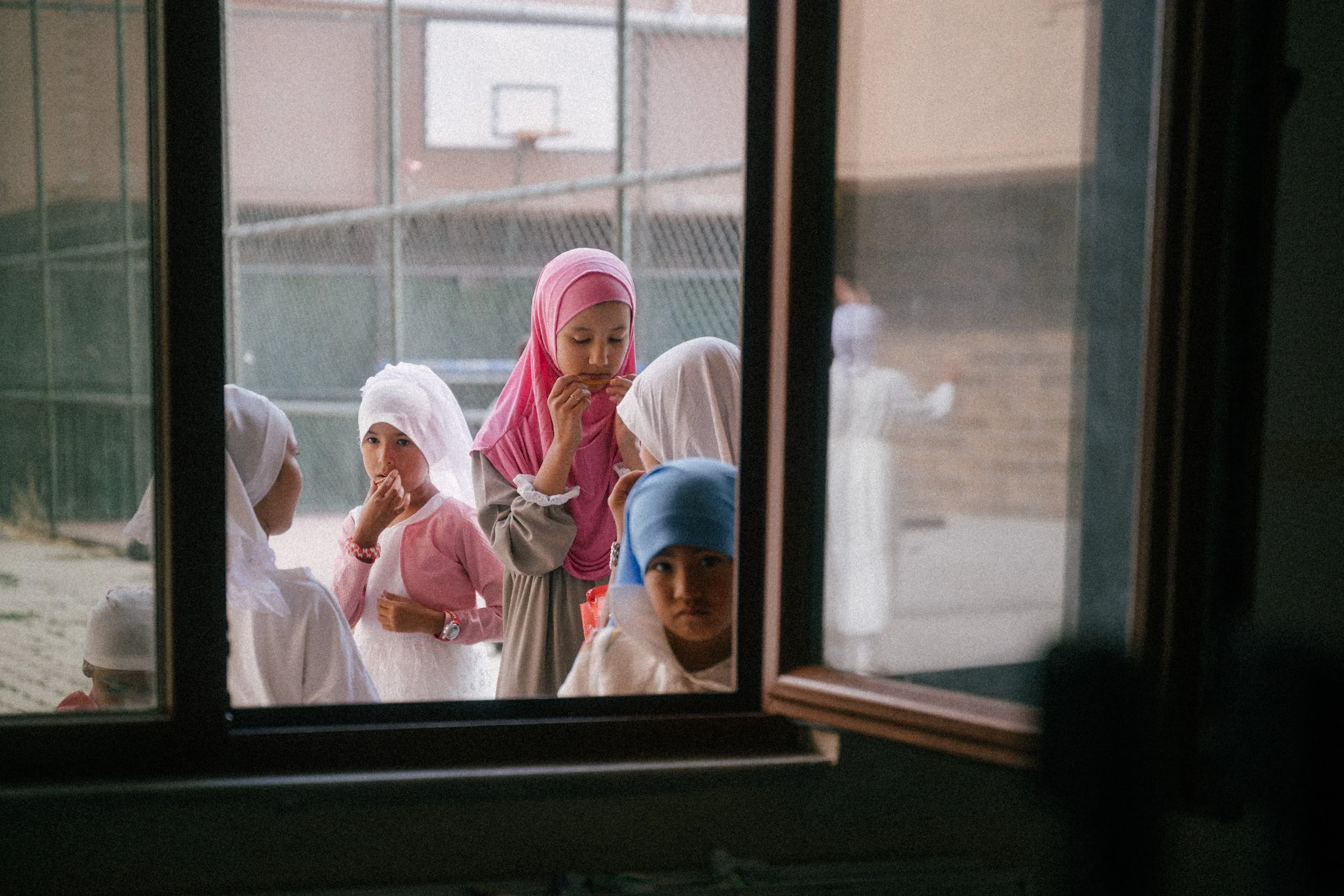 Children wearing hijabs standing outside a school window, some eating snacks and looking inside.