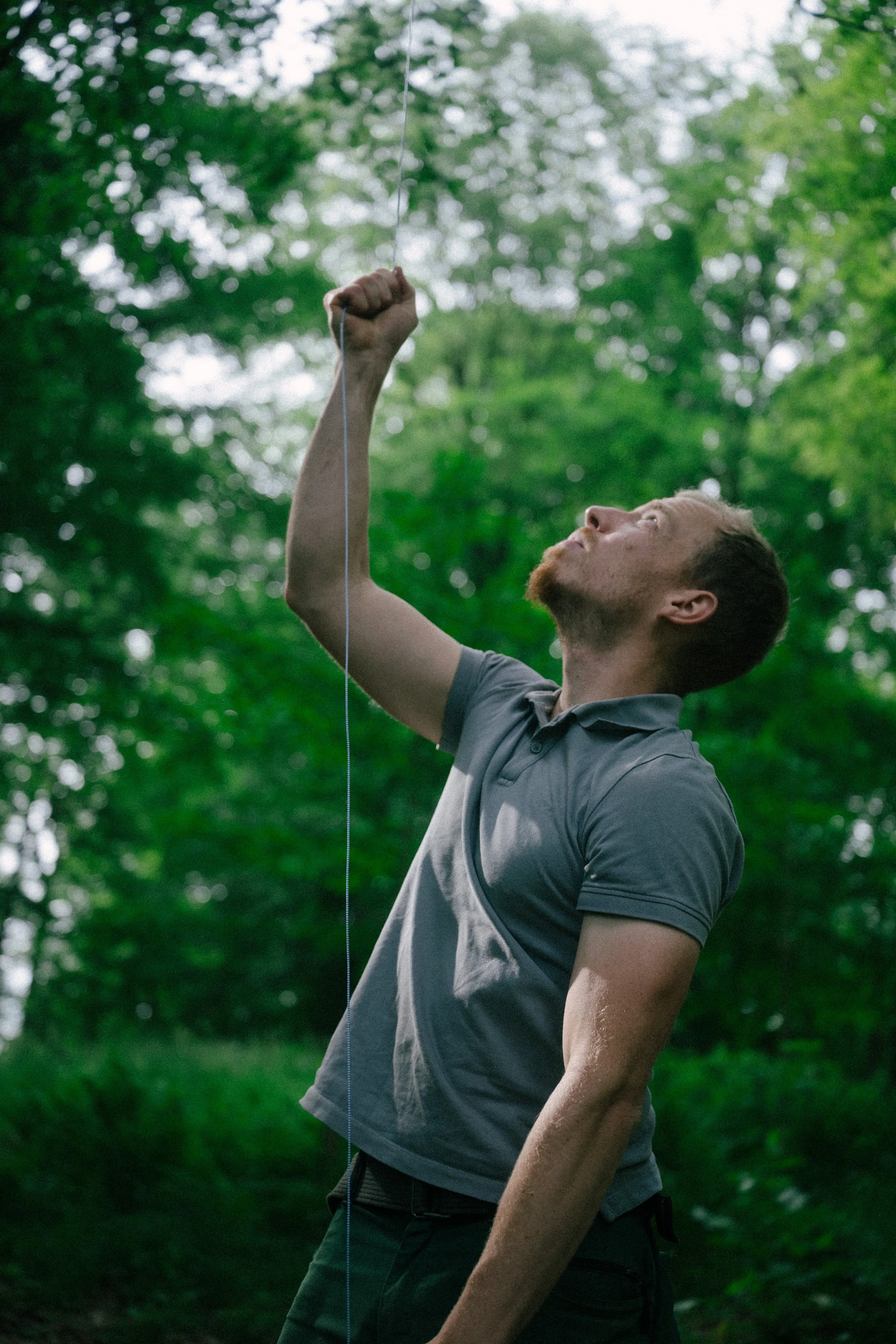 A man in a gray polo shirt and black pants standing outdoors, holding an upward-facing fist with his right hand, with a wooded background of green trees.
