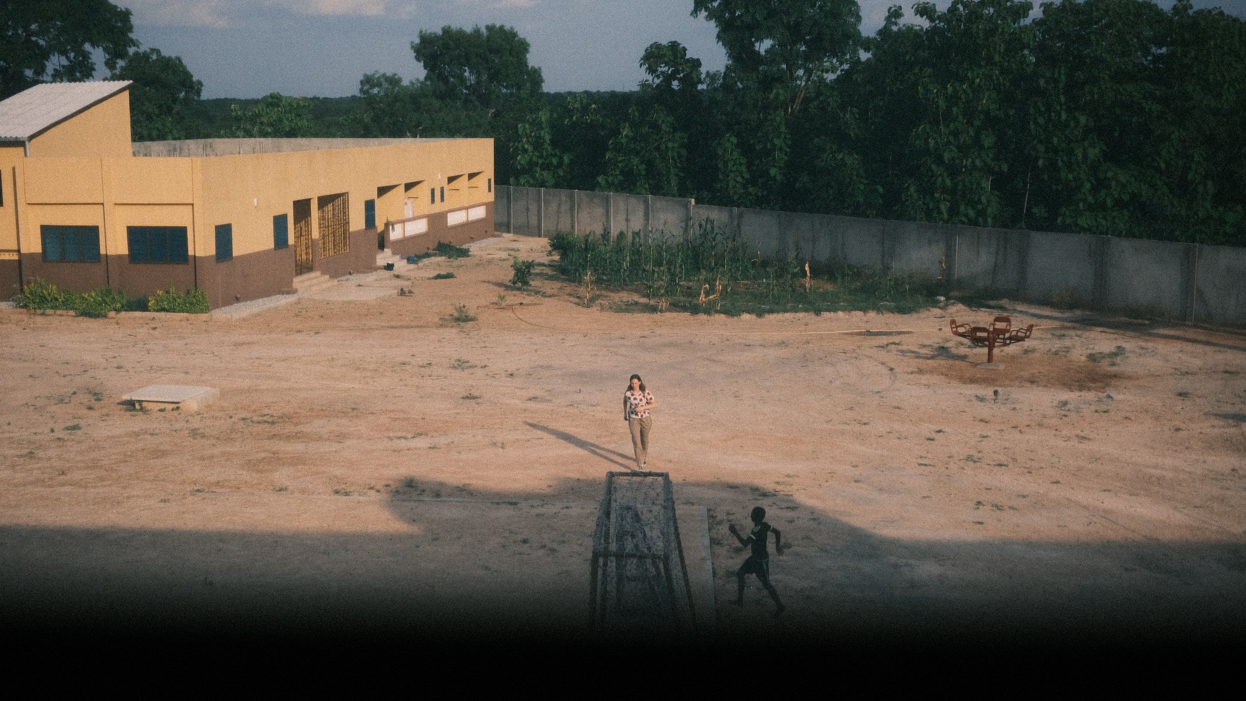 A woman stands on a court, preparing to hit a volleyball, with a young boy running towards her. There is a basketball hoop nearby, and a building with windows and plants in the background, surrounded by a concrete fence.