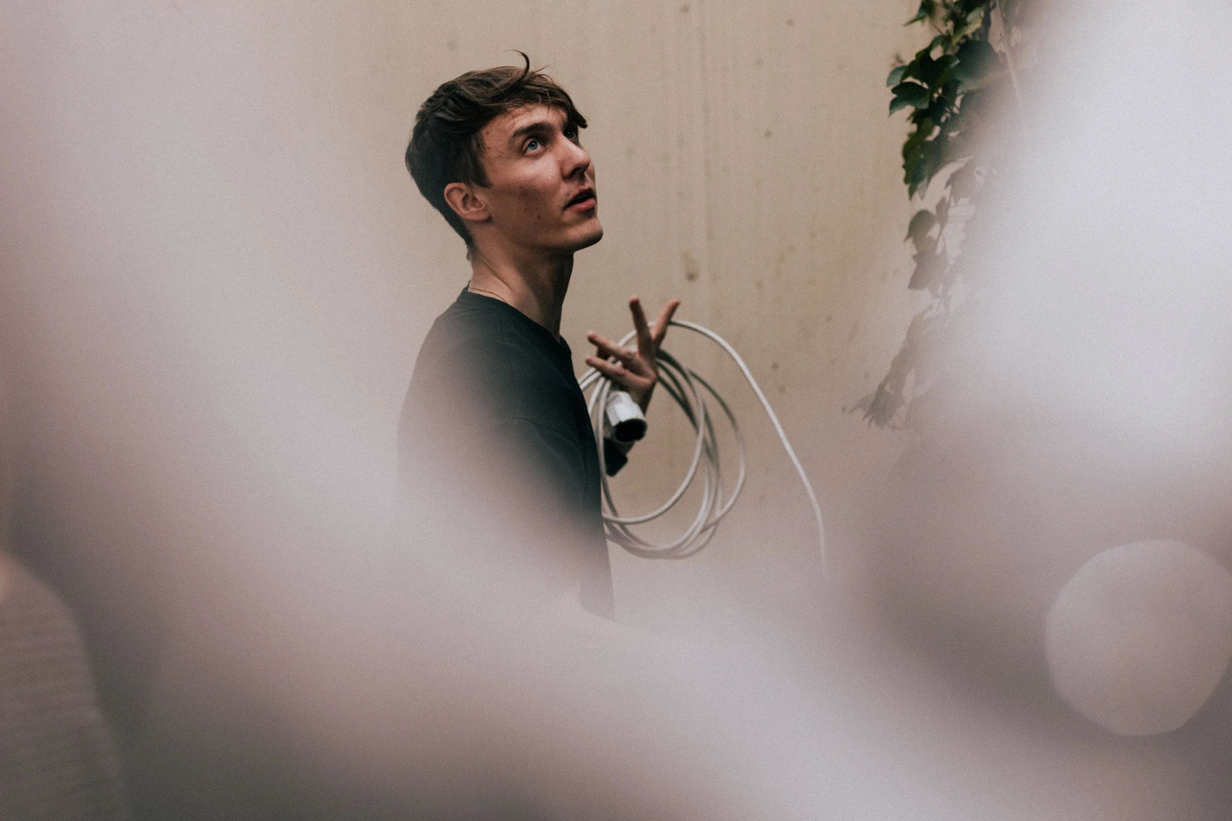 A young man with dark hair and fair skin looks upward while holding a bundle of white cables over his shoulder, standing against a beige wall with some greenery on the right side.