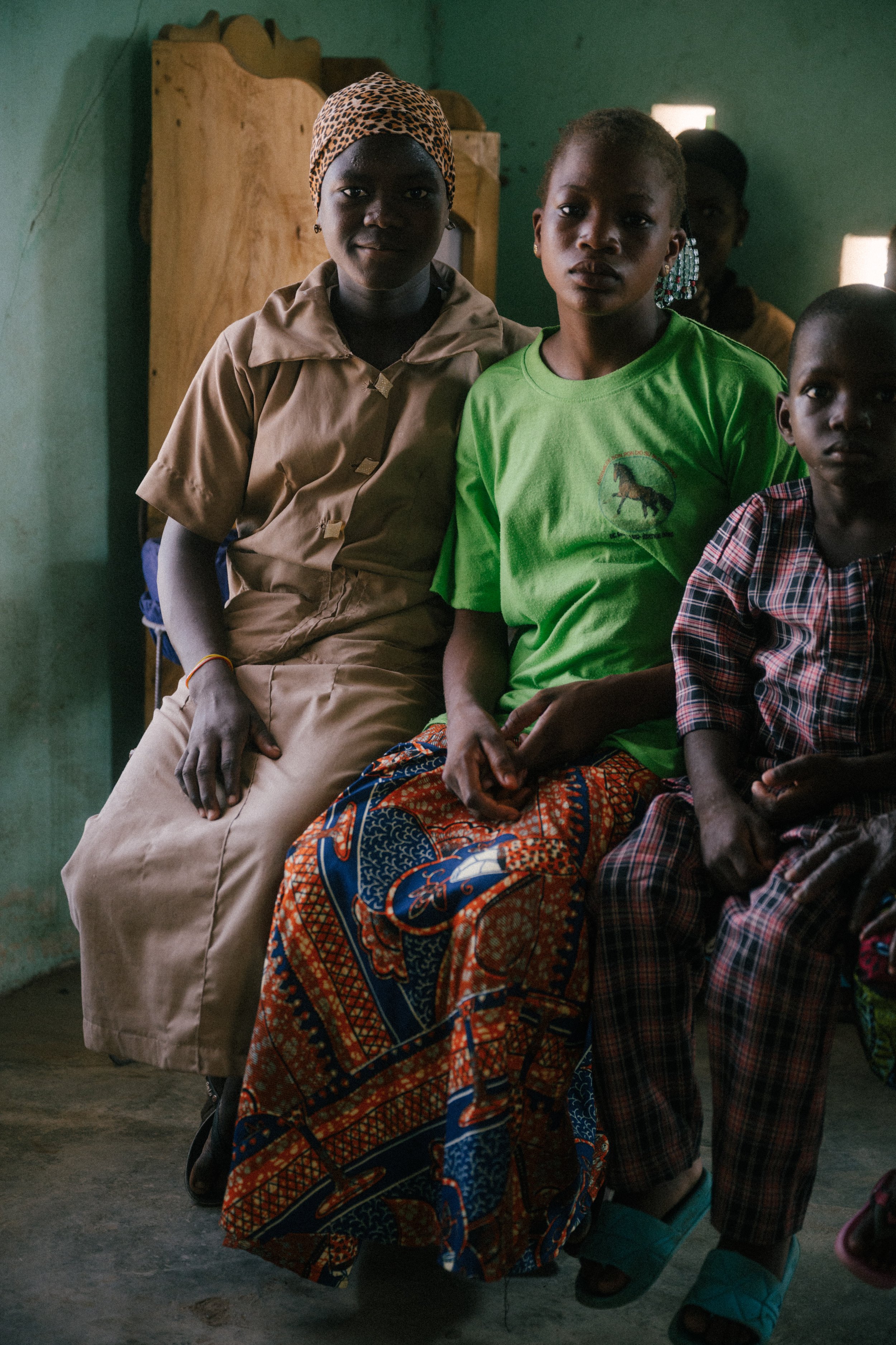 Three women and a young girl sitting on a bench inside a room with green walls, wearing casual clothes.