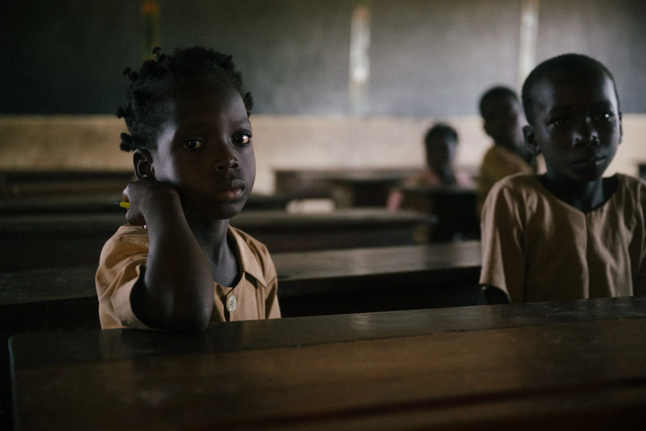 Children sitting in a classroom with wooden desks in a dimly lit room, looking towards the camera.