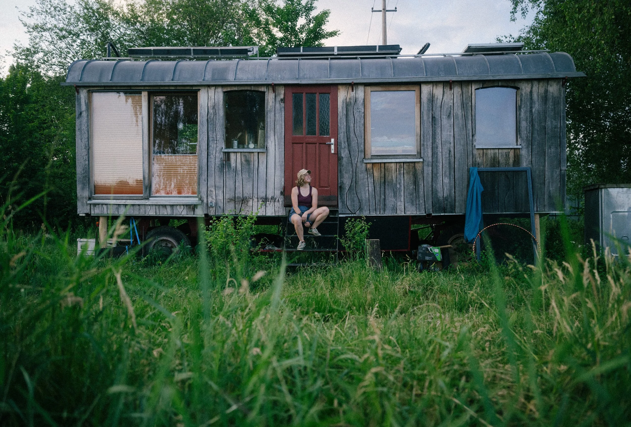 A rustic tiny house on wheels with weathered wood siding, surrounded by greenery, with a woman sitting on steps outside, wearing a hat and casual clothing.