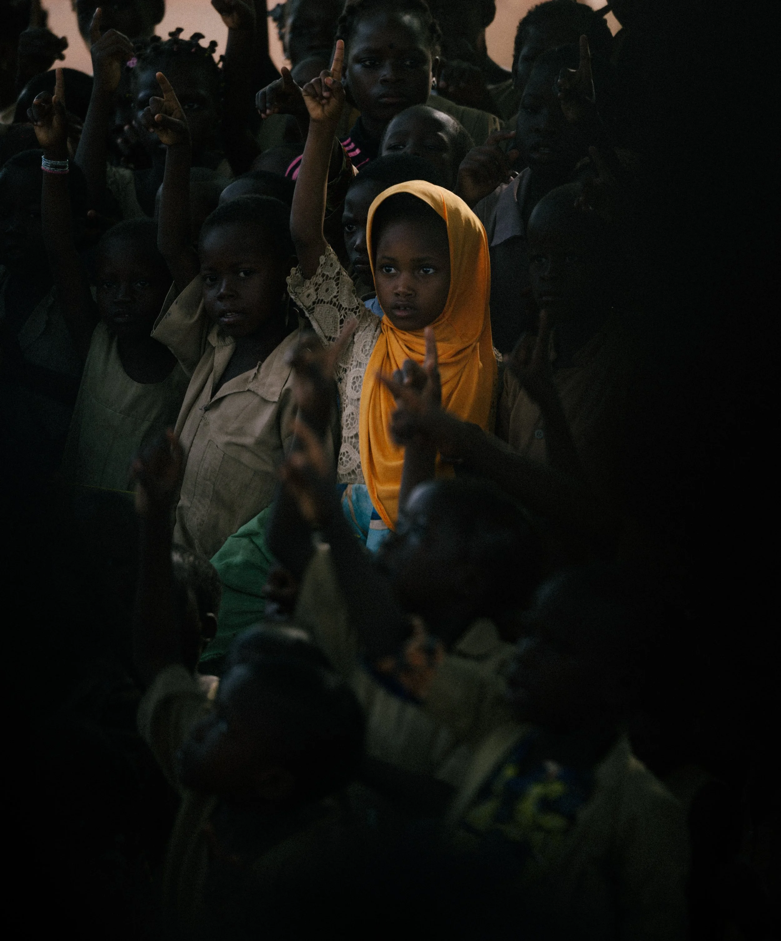Group of children gathered together, some raising their hands, with one girl wearing a yellow headscarf and clothing, in a dark setting.
