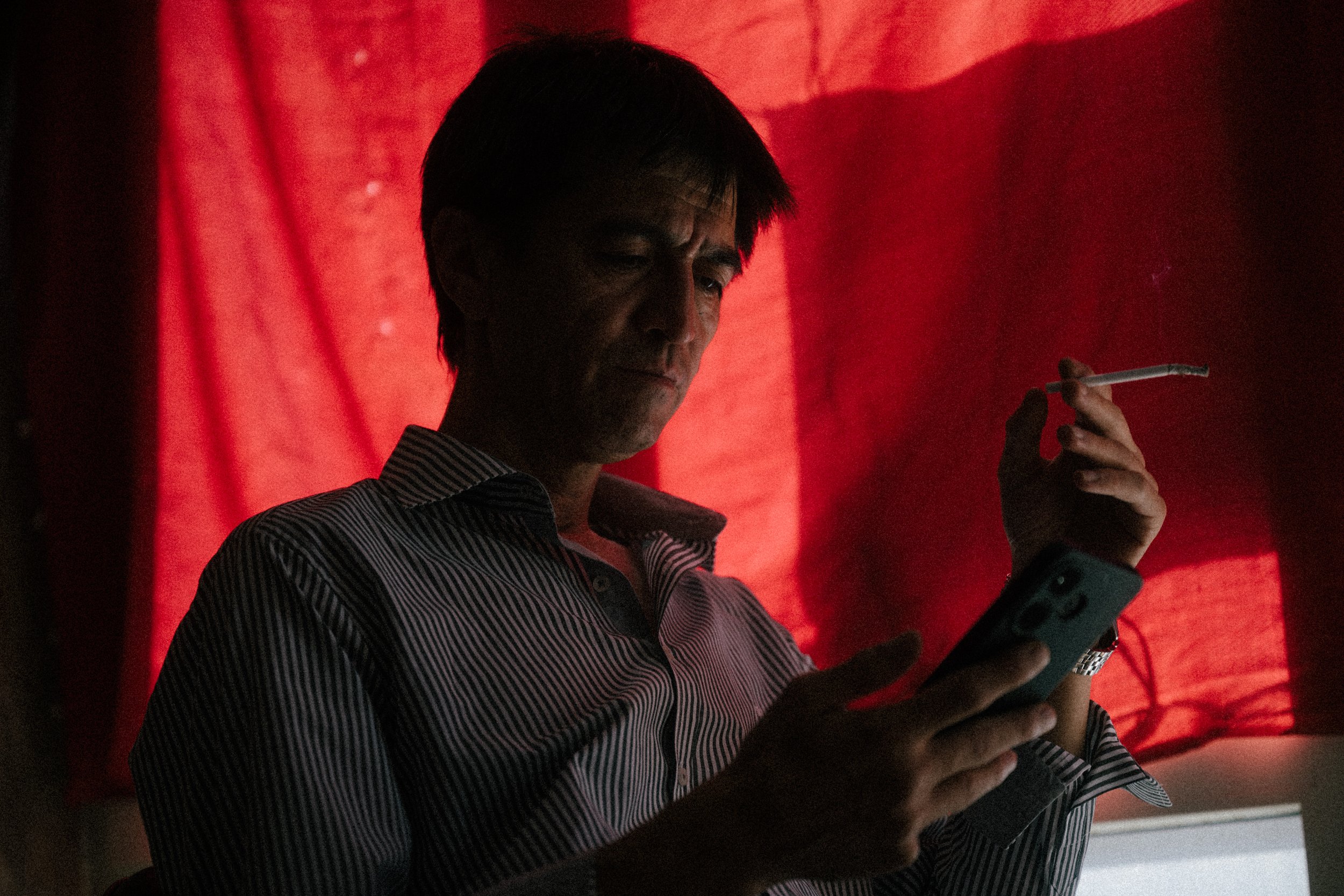 A man with dark hair wearing a striped shirt, holding a smartphone in one hand and a cigarette in the other, standing in front of a red curtain, with shadows cast on his face.