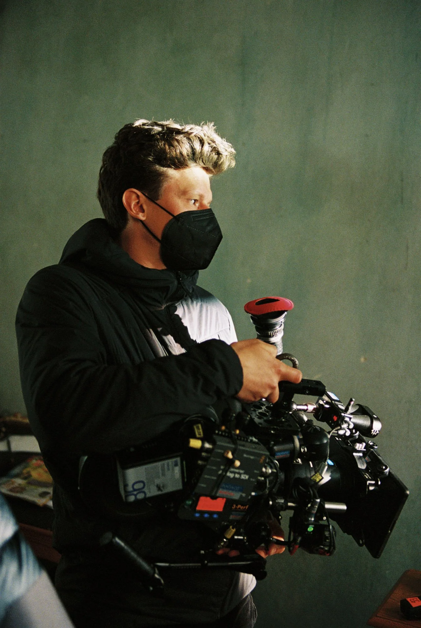 A young man wearing a black face mask and black jacket holding a professional video camera.
