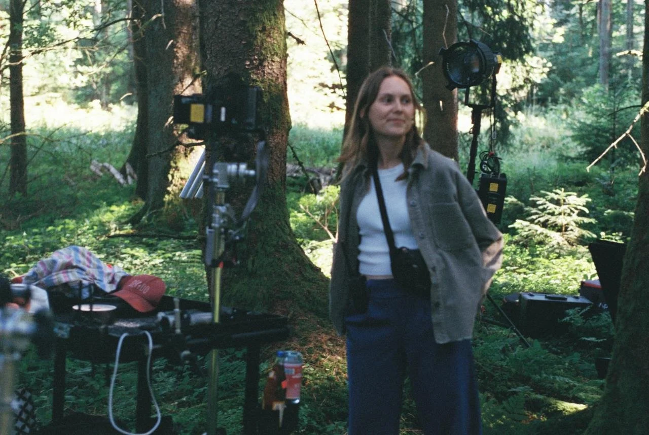 A woman standing in a forest surrounded by film equipment, including lights and cameras, indicating a film or photography shoot.
