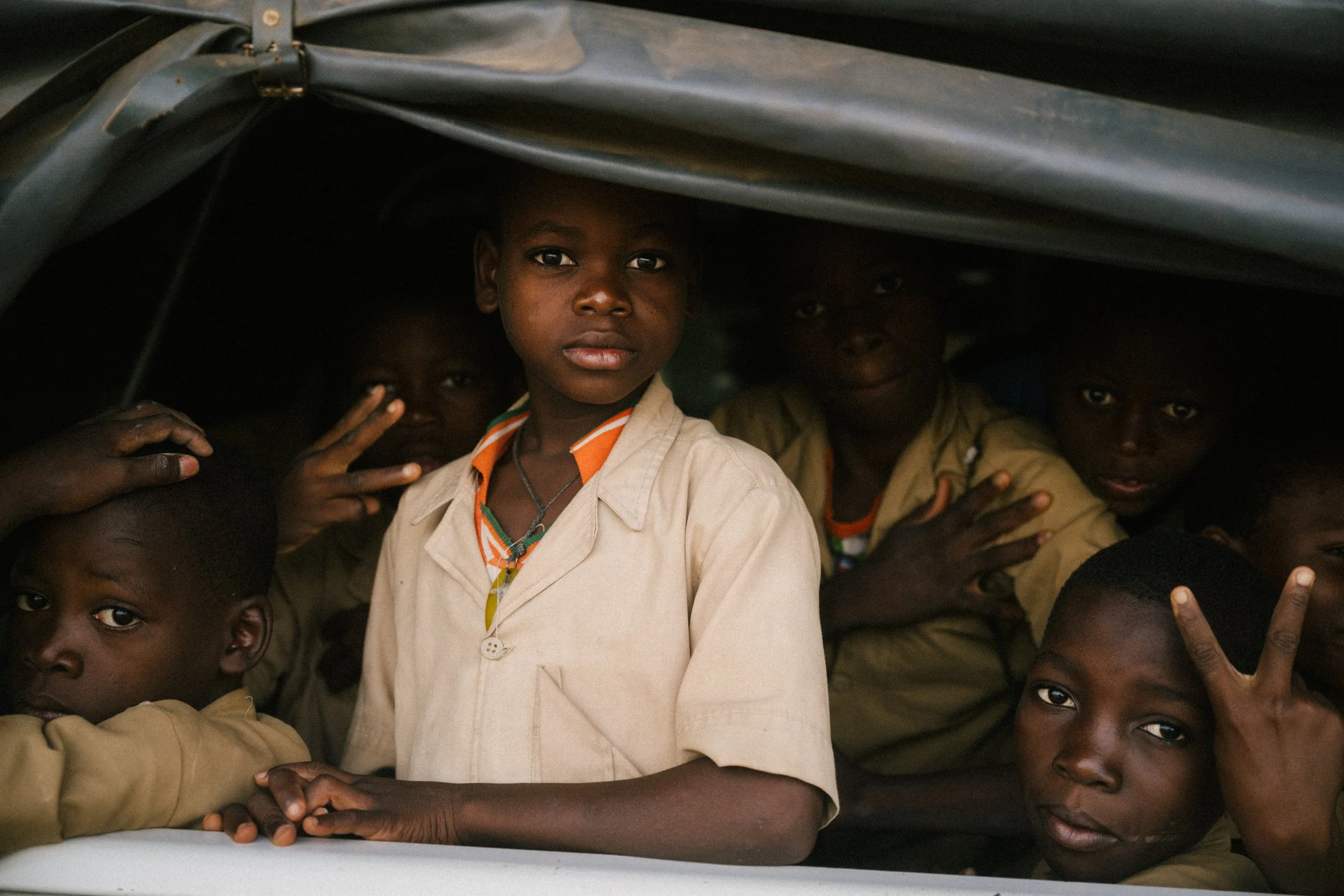 Group of young African children inside a vehicle, some making peace signs, with one girl in the center looking out.