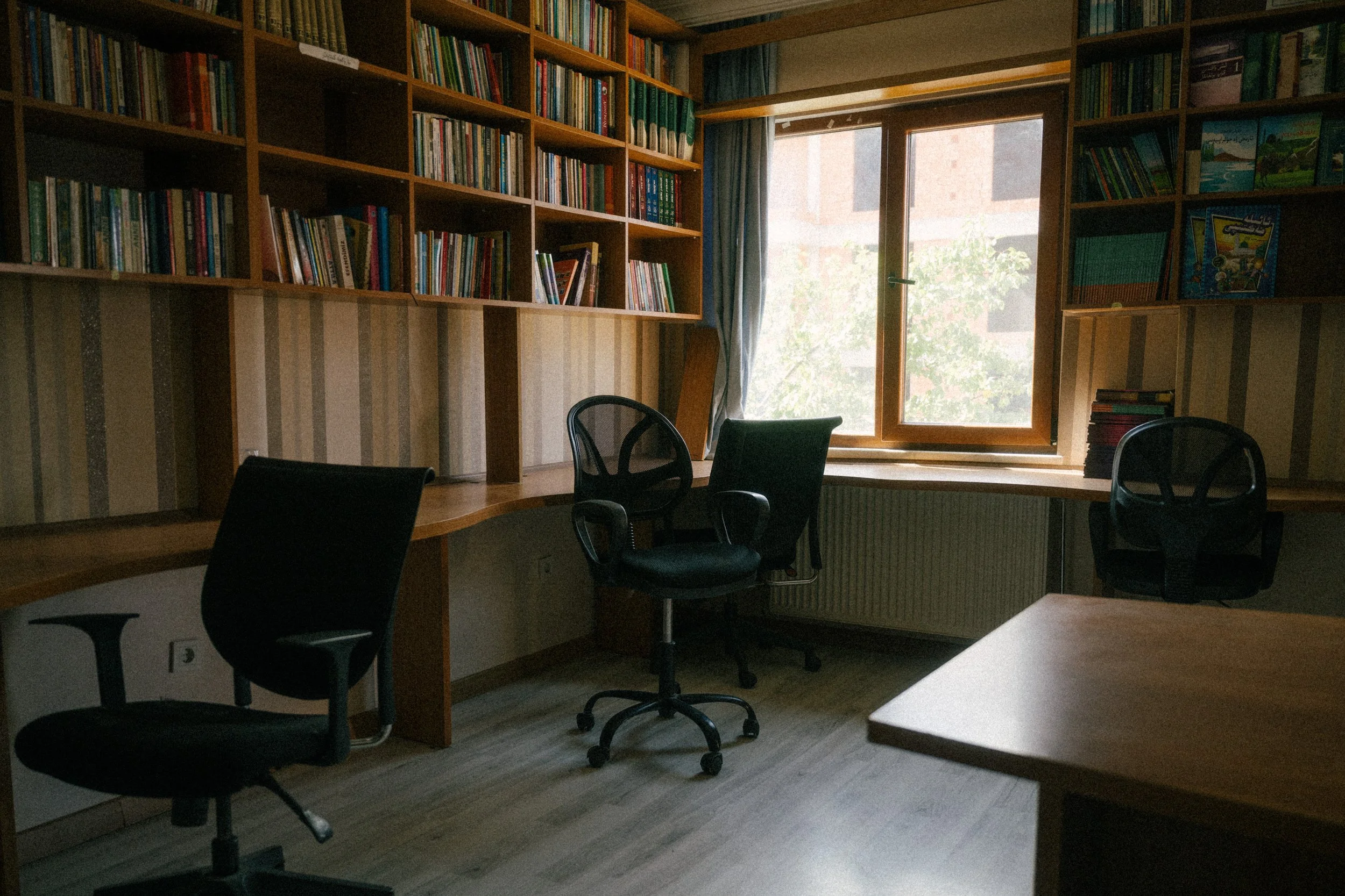 A cozy home office with built-in wooden bookshelves filled with colorful books, a large window letting in natural light, a curved desk, and three black office chairs.