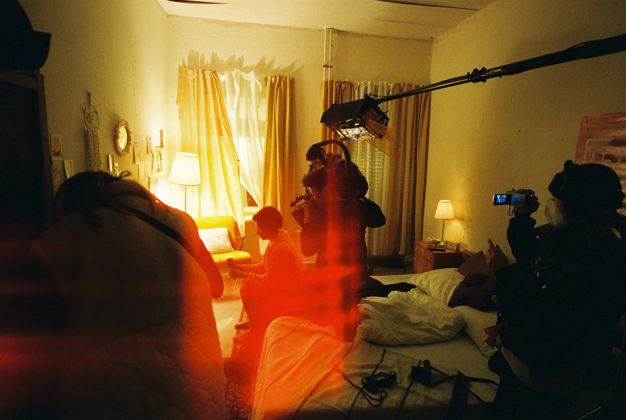 A behind-the-scenes shot of a film or video production in a cozy living room. There is a woman sitting on a yellow sofa, a crew member holding a large camera rig, another person filming with a smartphone, a soft yellow light, and a large light source