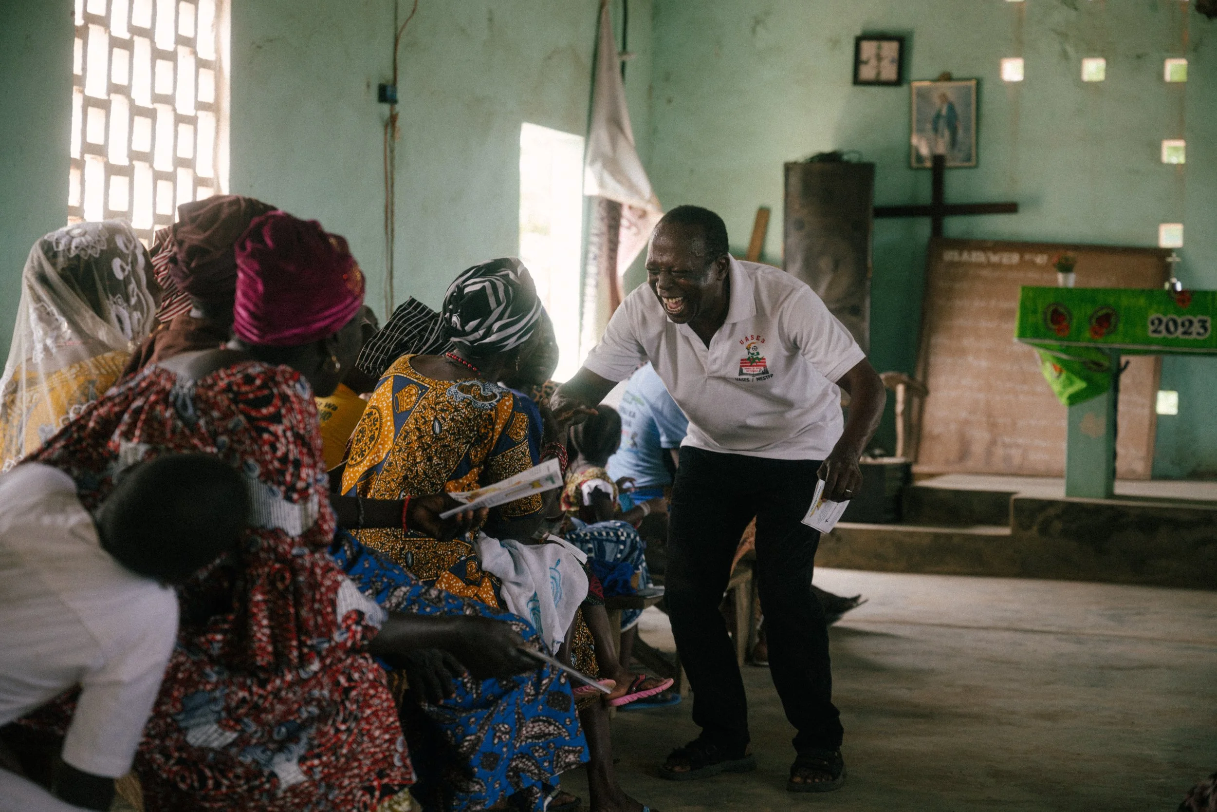 A man smiling and engaging with a group of women seated in a rustic community room, with some women holding papers, in a setting with green walls and minimal decoration.