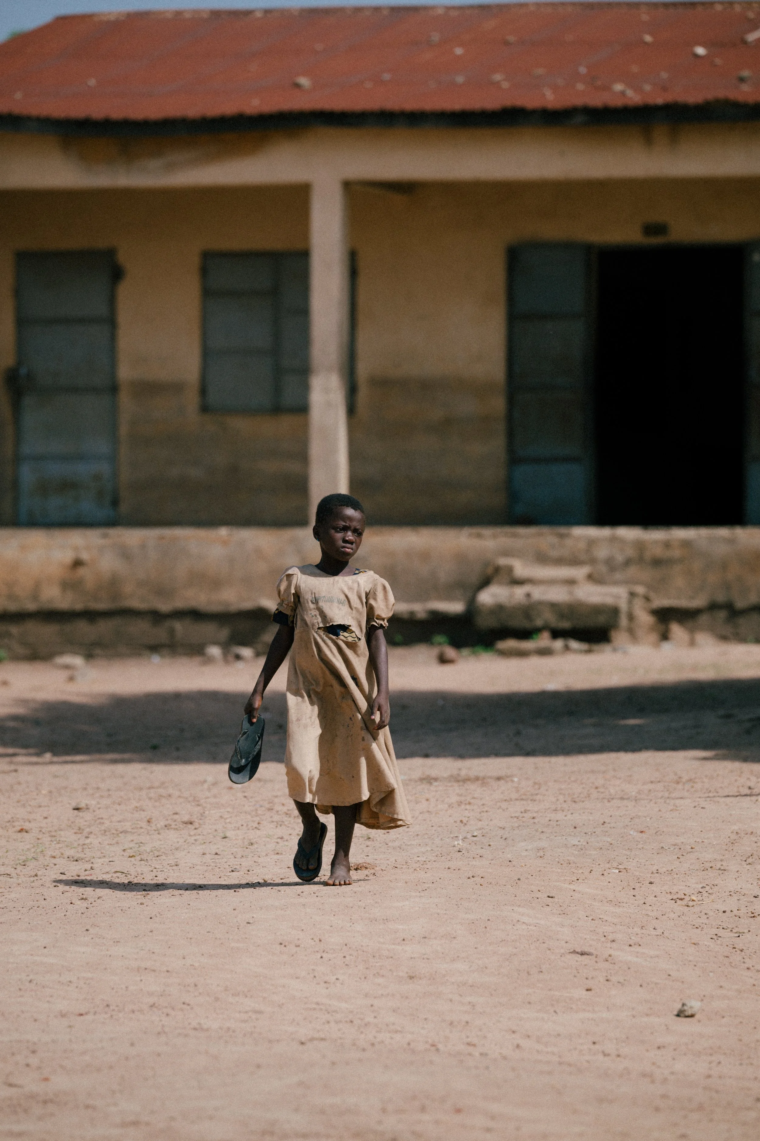 A young girl walking barefoot on a dirt street, holding a sandal in her hand, with a worn house in the background.