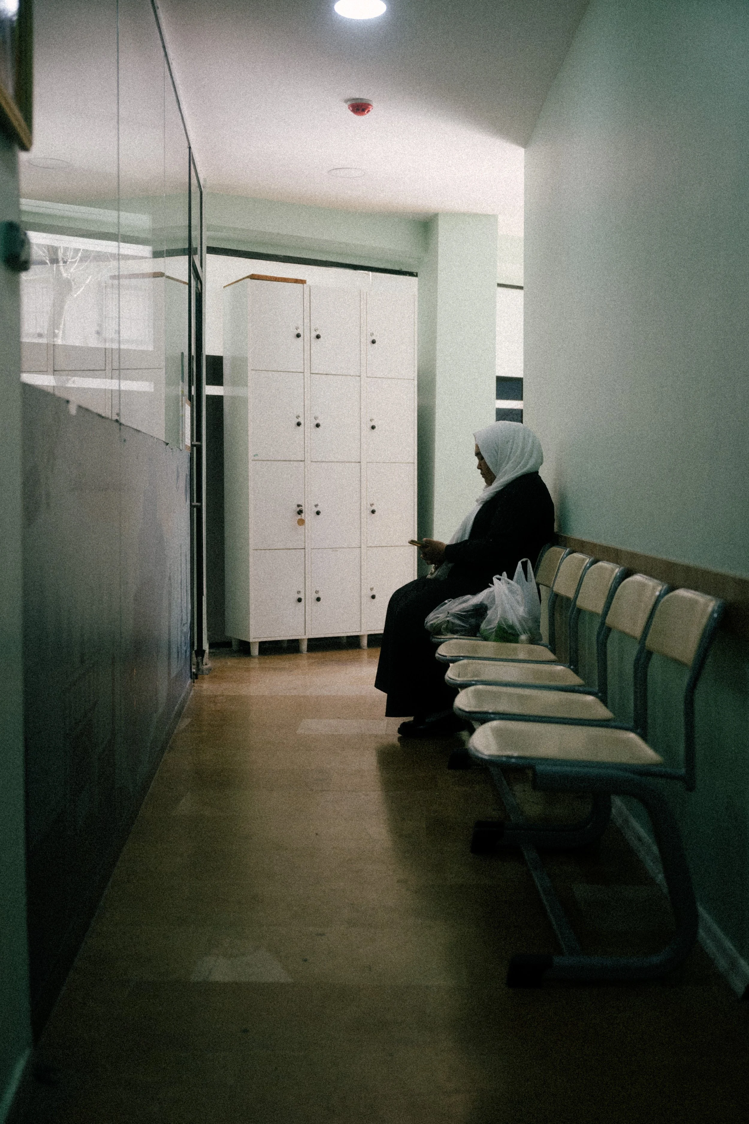 A woman wearing a hijab sitting alone on a row of empty chairs in a waiting area, looking at her phone, with lockers behind her.