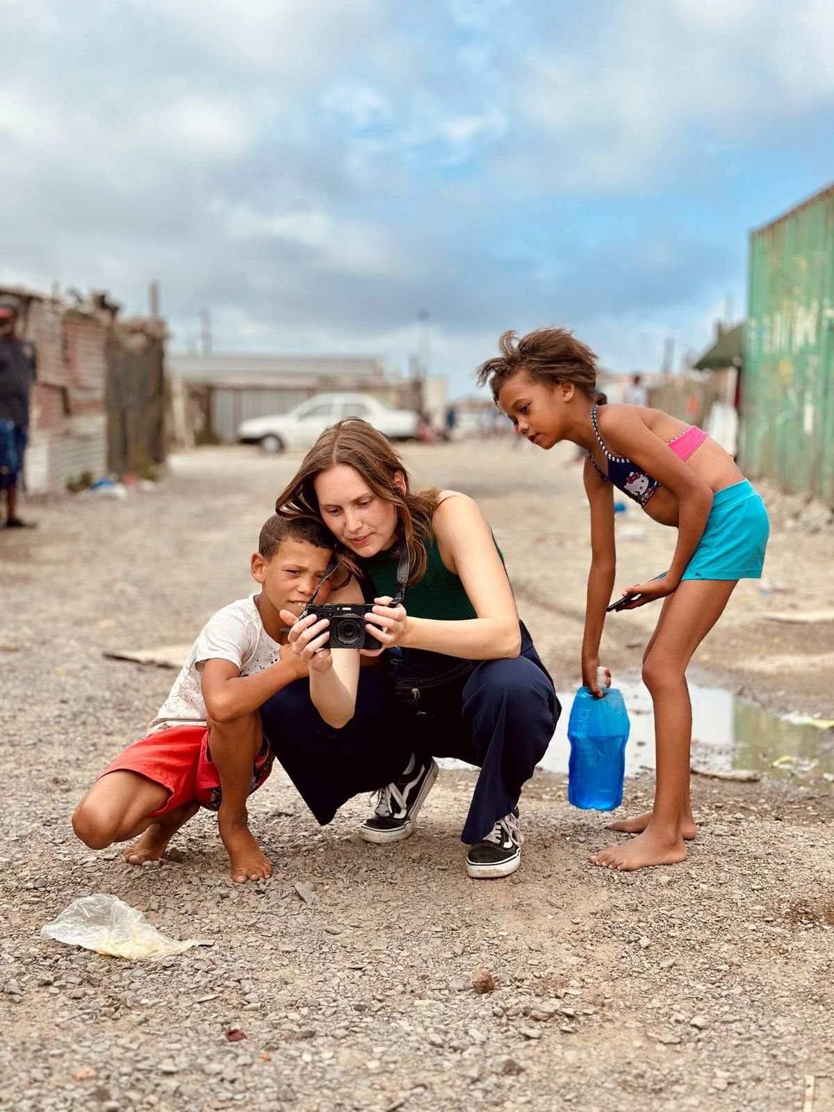 A woman and two children standing on a dirt road, looking at a camera the woman is holding. The girl is holding a plastic bottle, and the boy is crouched down in front of them, looking at the camera.