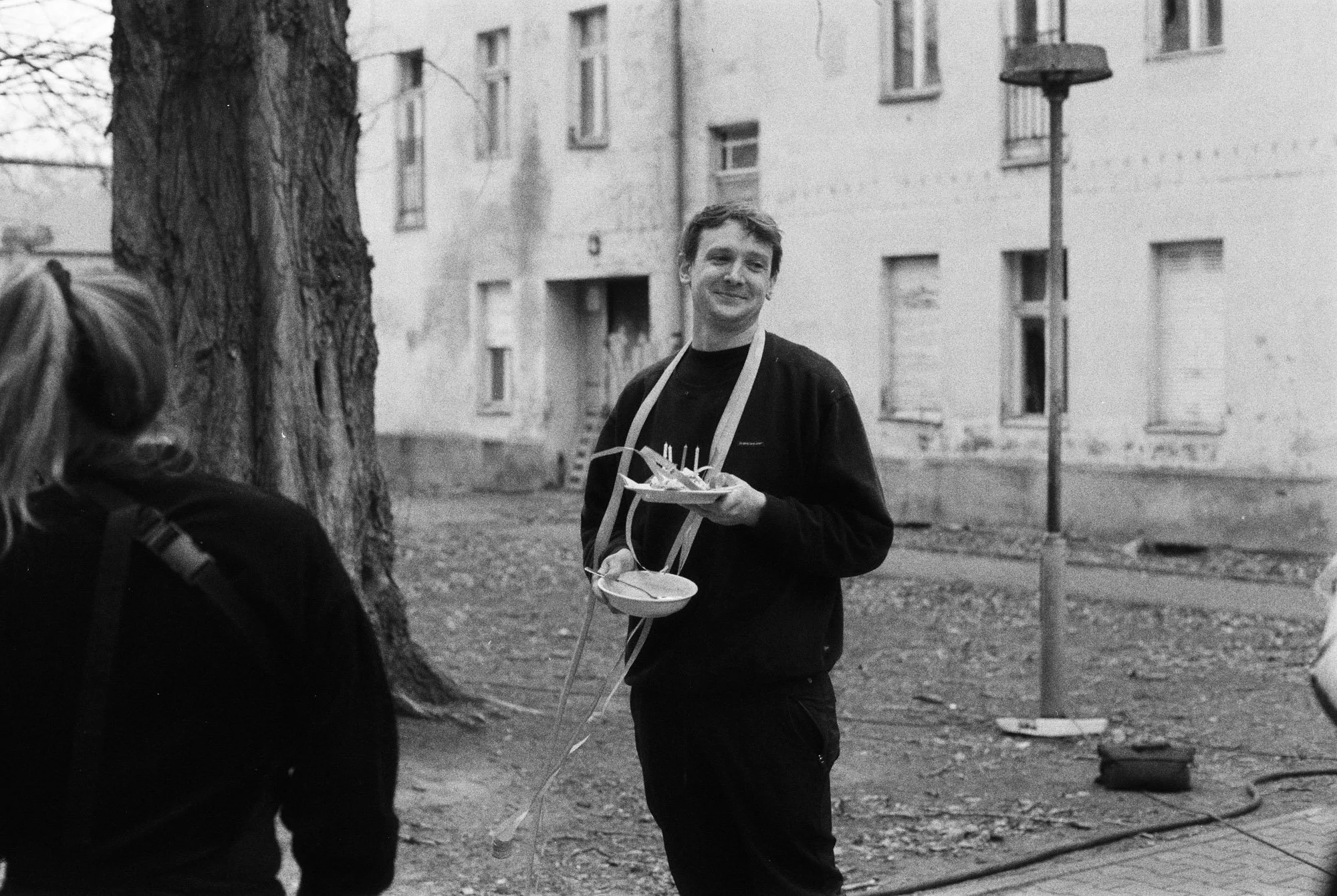 A young man stands outdoors near a large tree, holding a plate of food with a smile on his face. A woman with a backpack is partially visible in the foreground, and there is a streetlamp and residential building behind him.