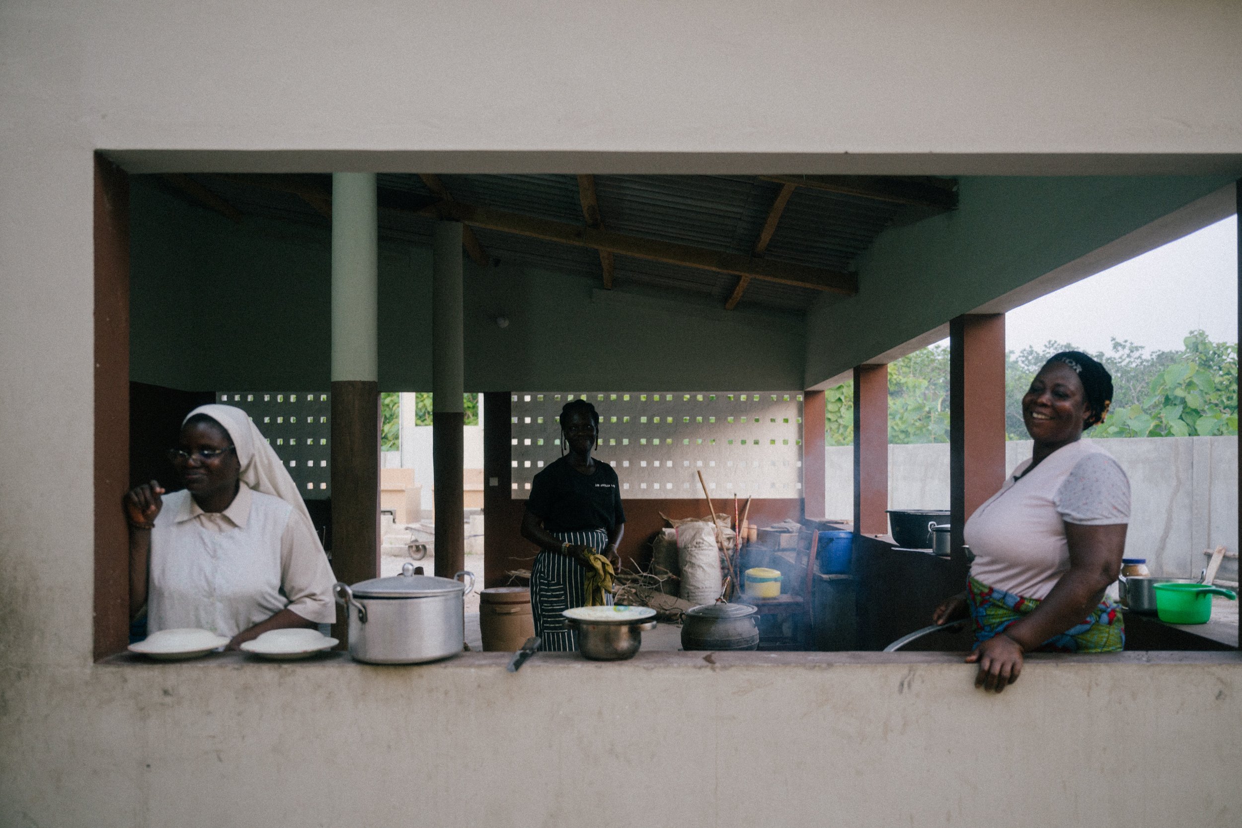 Three women cooking and smiling in an outdoor kitchen with large open window