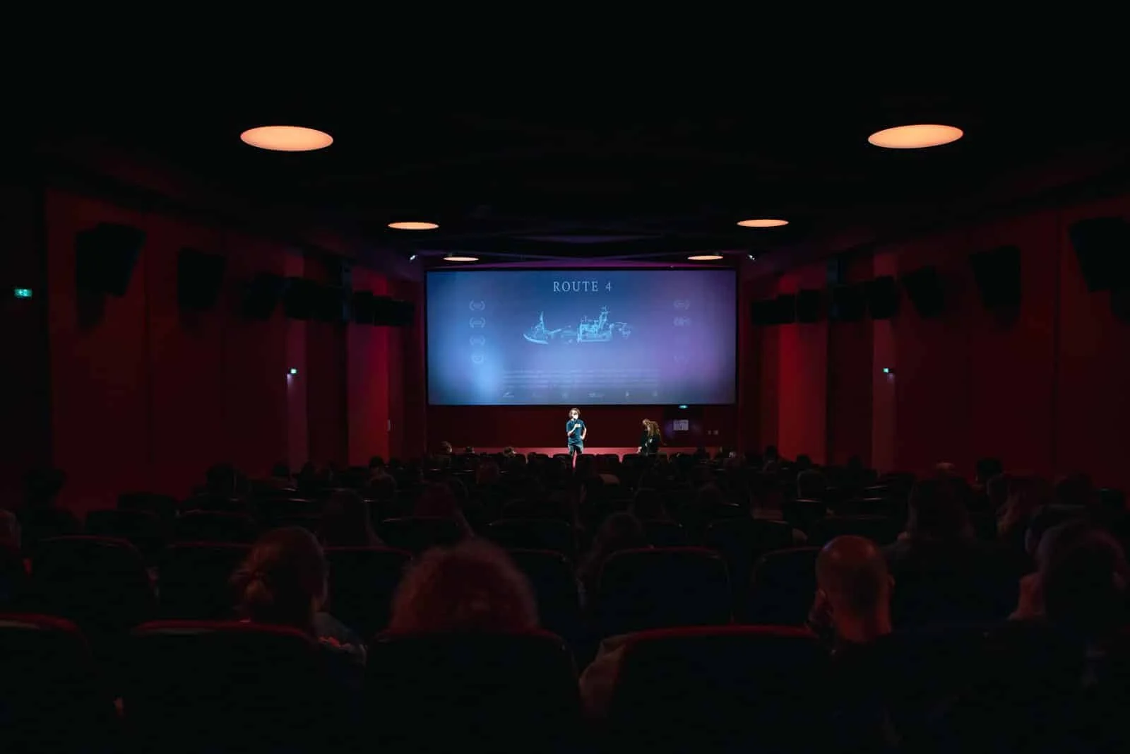 A movie theater with audience members watching a presentation on the large screen. The screen displays the words 'ROUTE 4' and a schematic diagram.