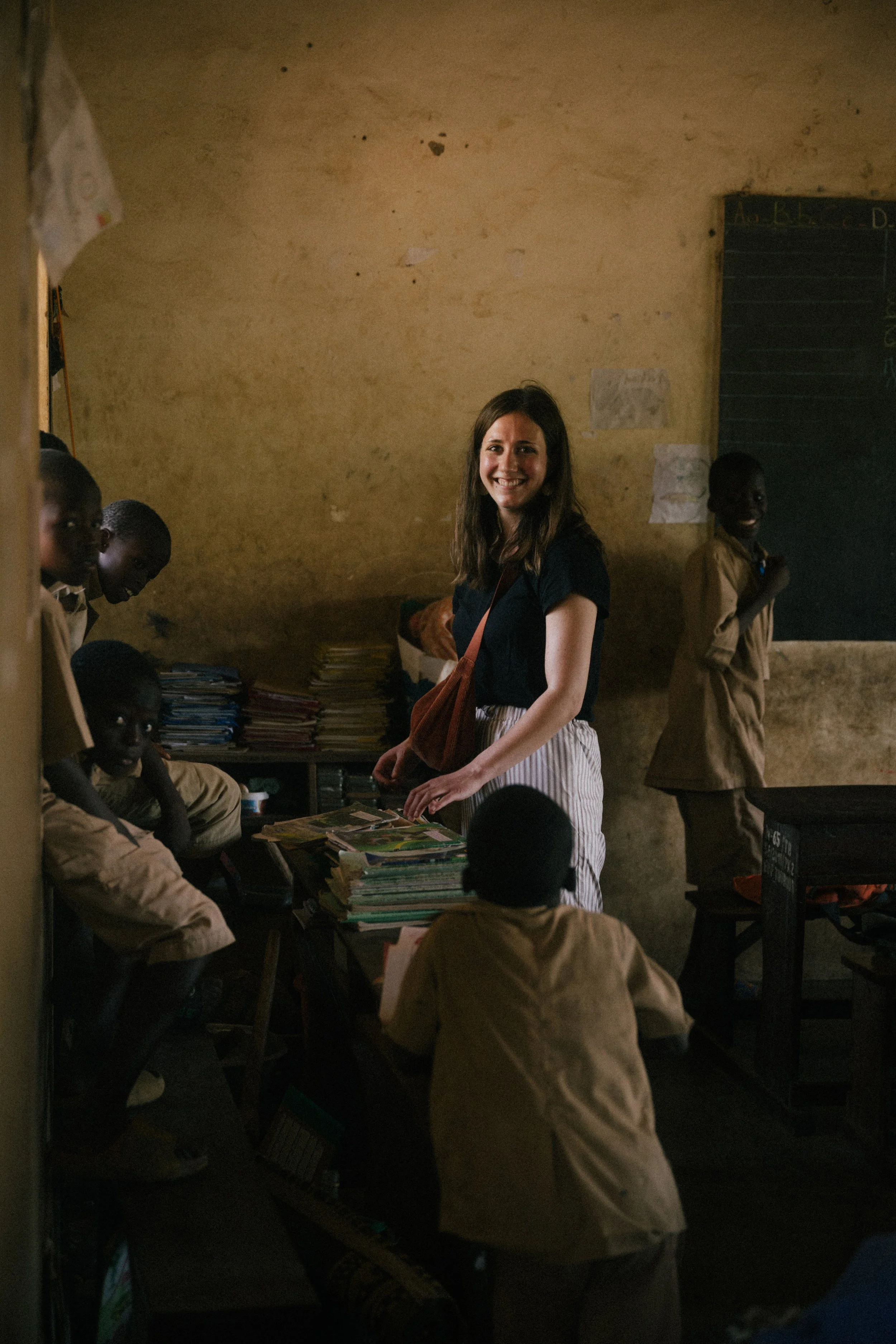A smiling young woman with brown hair stands in a classroom with dark yellow walls, surrounded by children in beige uniforms. She is looking at the camera, with books stacked on the table in front of her, and other students partially visible around h