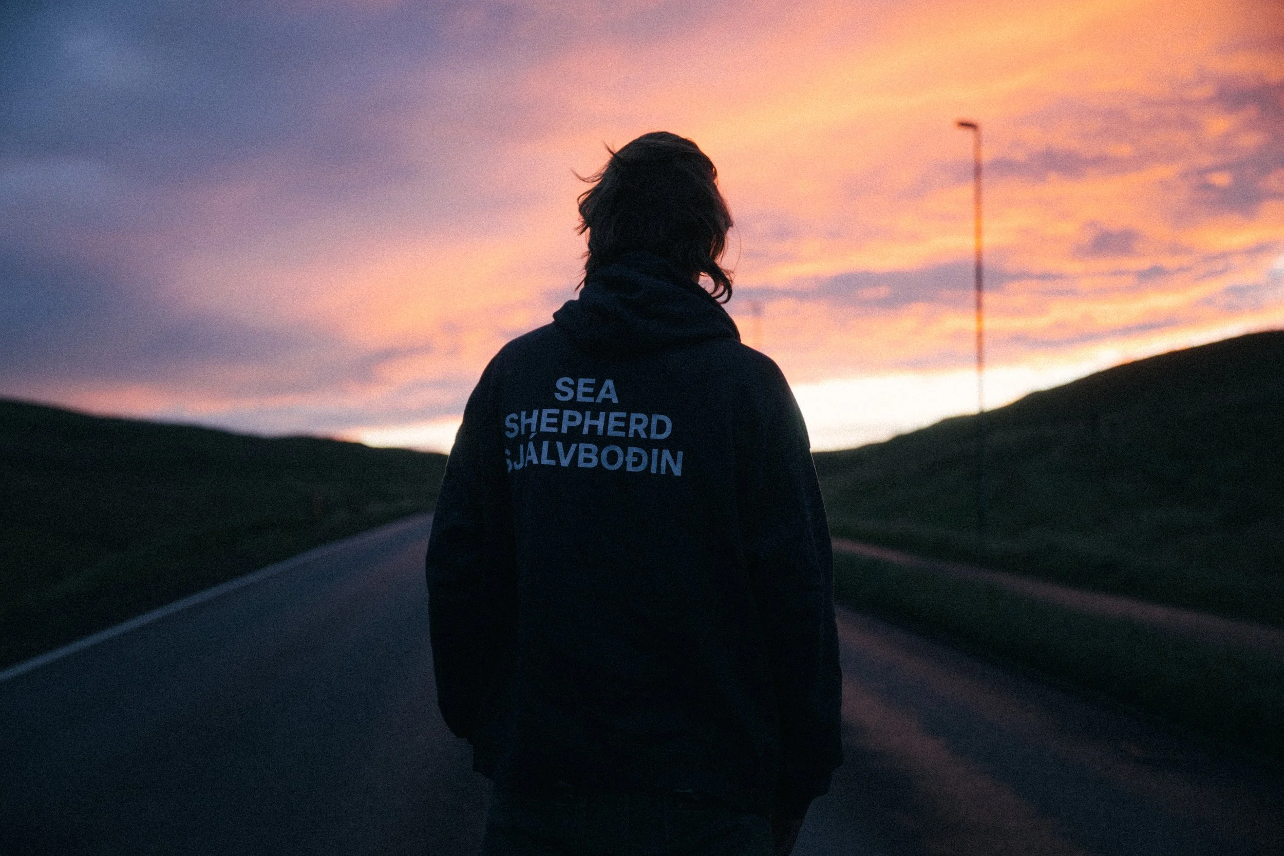 A person standing on a road at sunset, wearing a black hoodie with the words 'SEA SHEPHERD JÁLVBODIN' on the back.