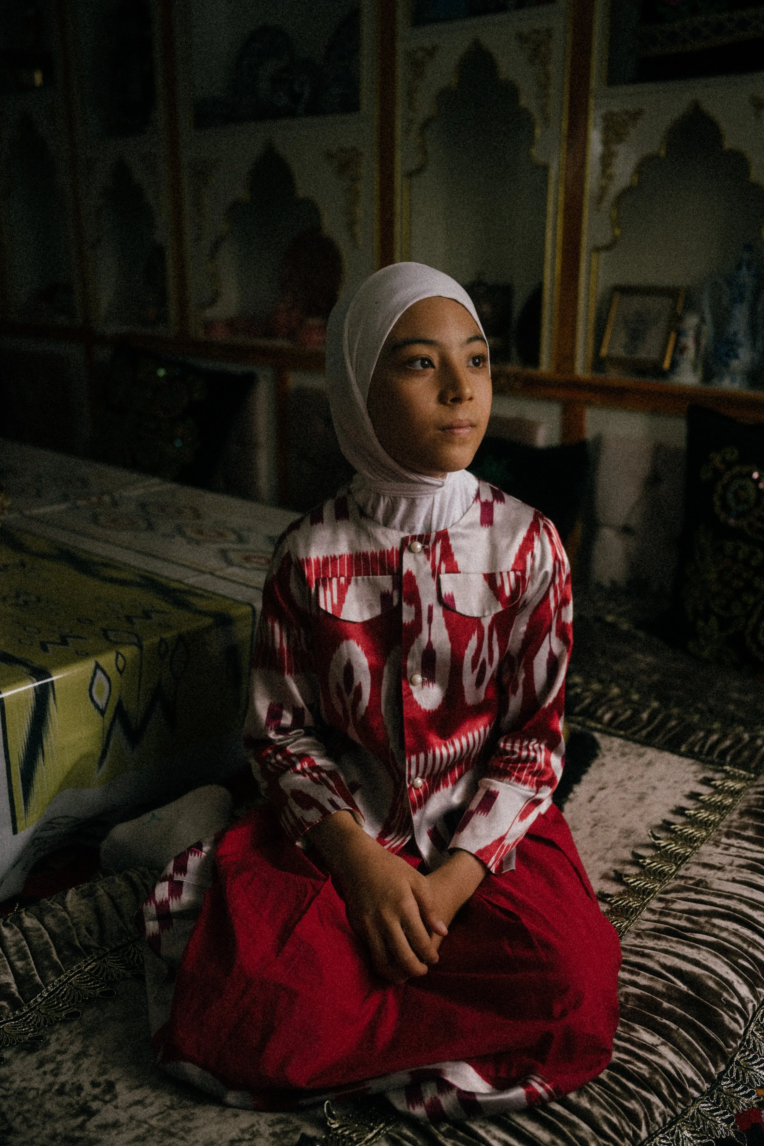A young girl in a white hijab and patterned red and white outfit sitting on a cushion in a dimly lit room with traditional decor.