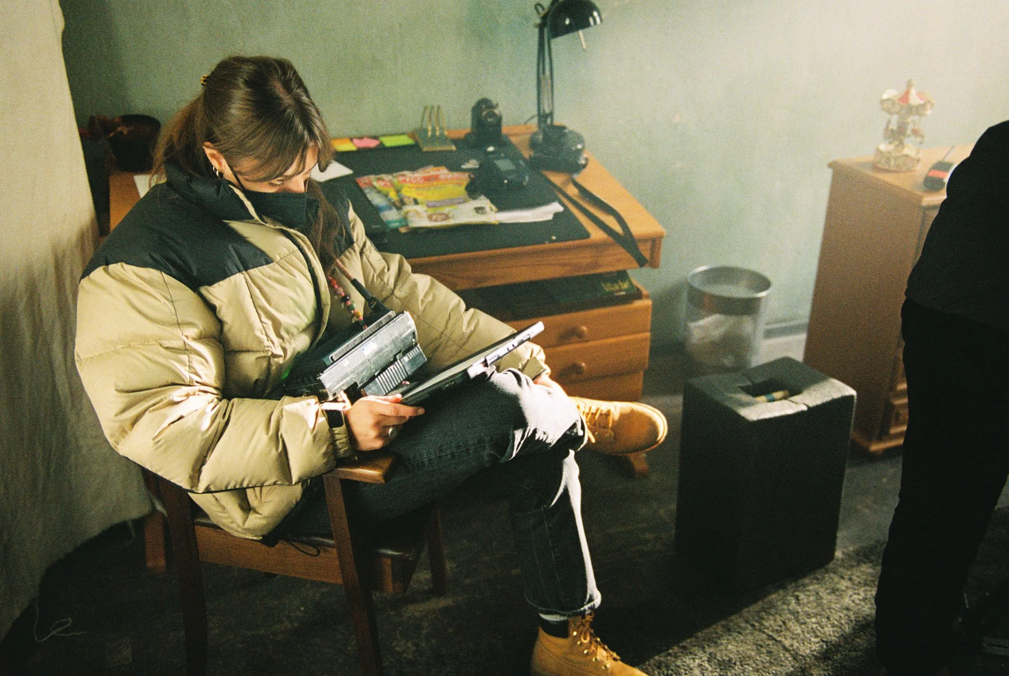 A woman sitting on a chair in a room, wearing a beige and black puffer jacket, yellow boots, and a mask, looking at her phone. There is a desk behind her with various items, including magazines, bags, and electronics. To her right, there is a trash c