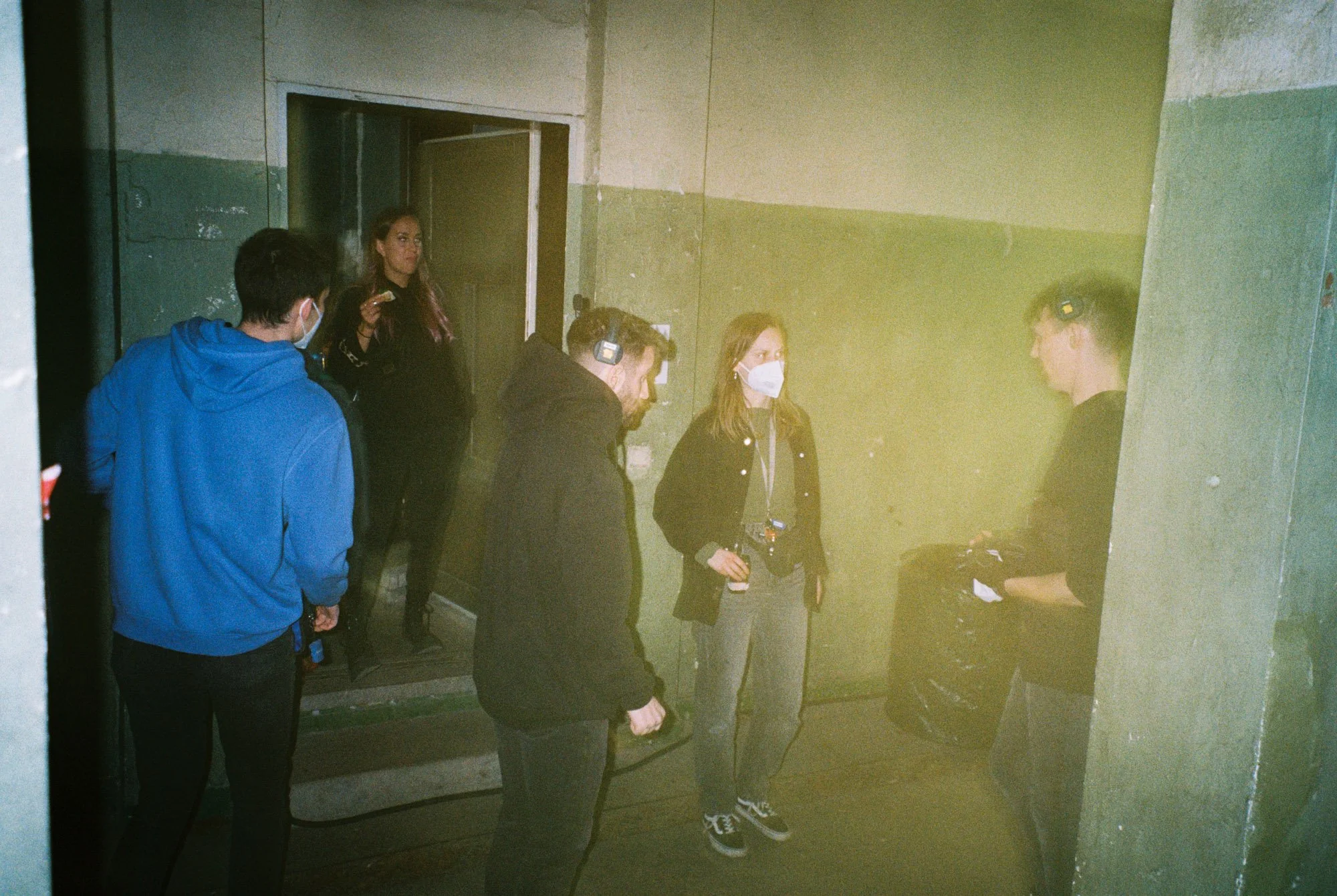 A group of five people, some wearing face masks, standing in a dimly lit, industrial-looking room with concrete walls. They appear to be engaged in a discussion or listening to a person who is speaking and holding items. One person is taking notes or