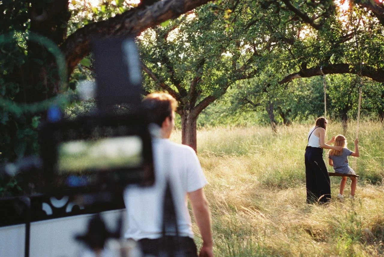A woman pushes a young girl on a swing in a grassy field shaded by trees during daylight, with a man operating a camera in the foreground.