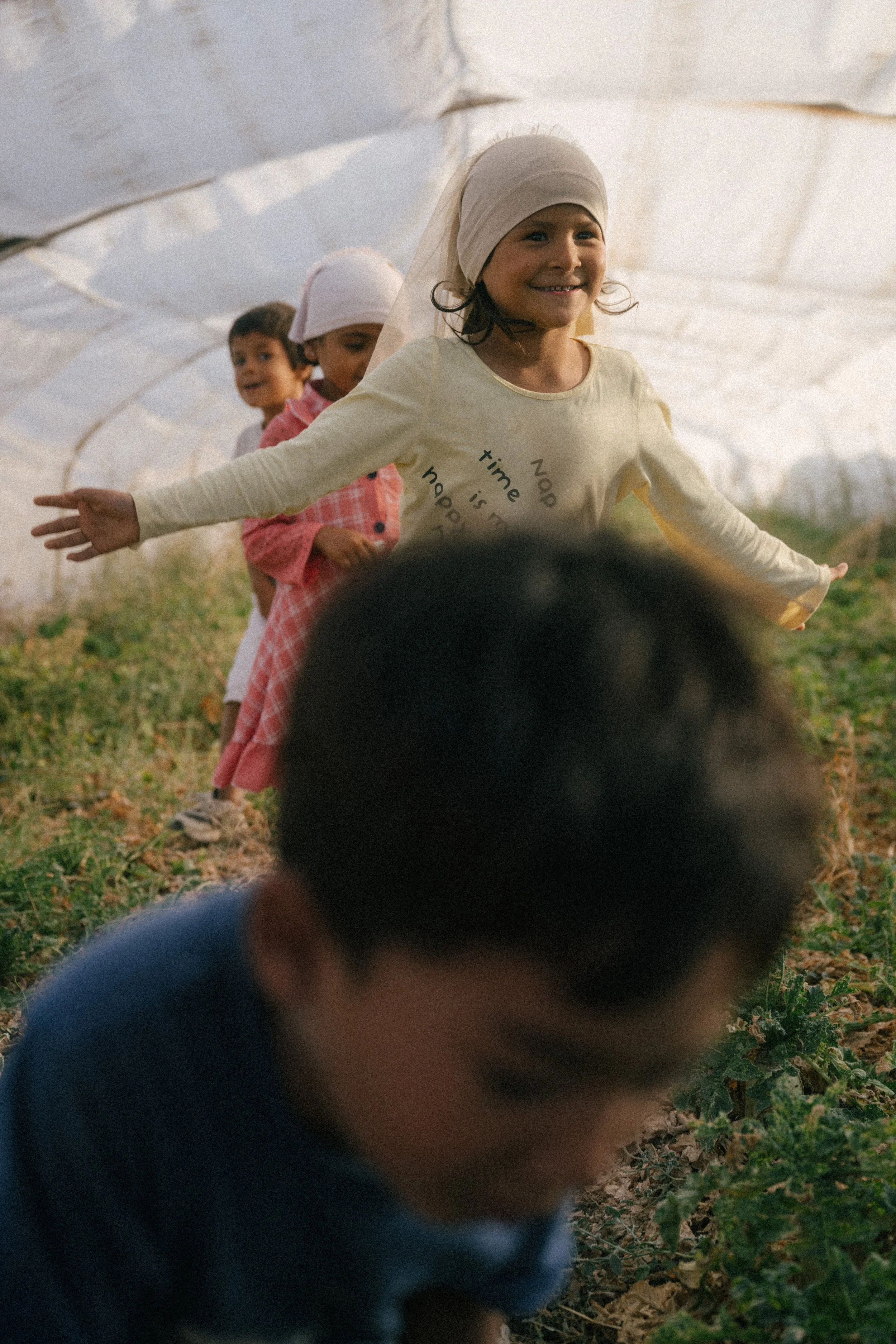 A group of four children standing in a greenhouse, with one child in the foreground and three children smiling in the background, with natural light coming through the plastic covering of the greenhouse.