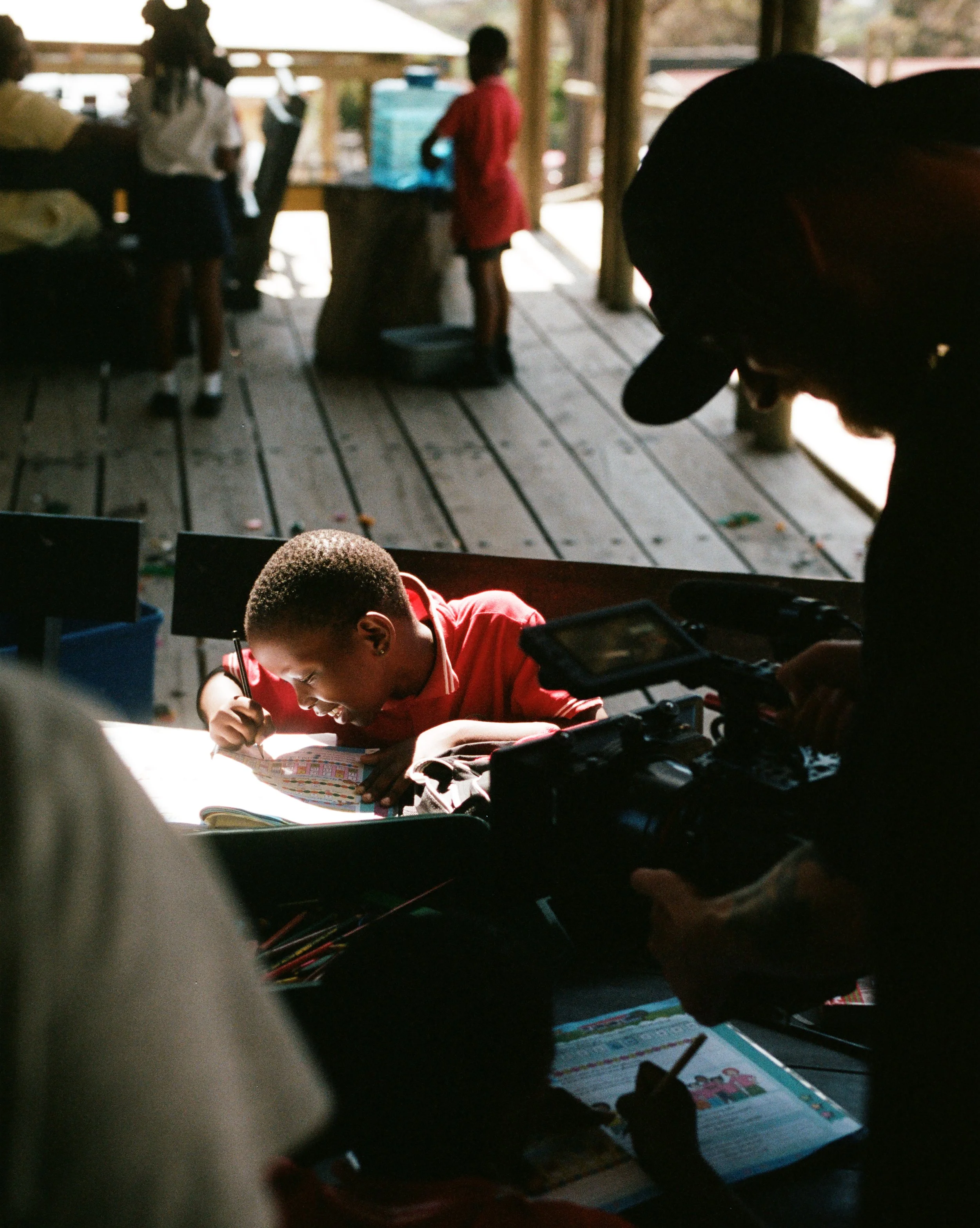 A boy in a red shirt is writing in a notebook with a smile, while a person with a camera films him. Other children are in the background on a wooden deck.